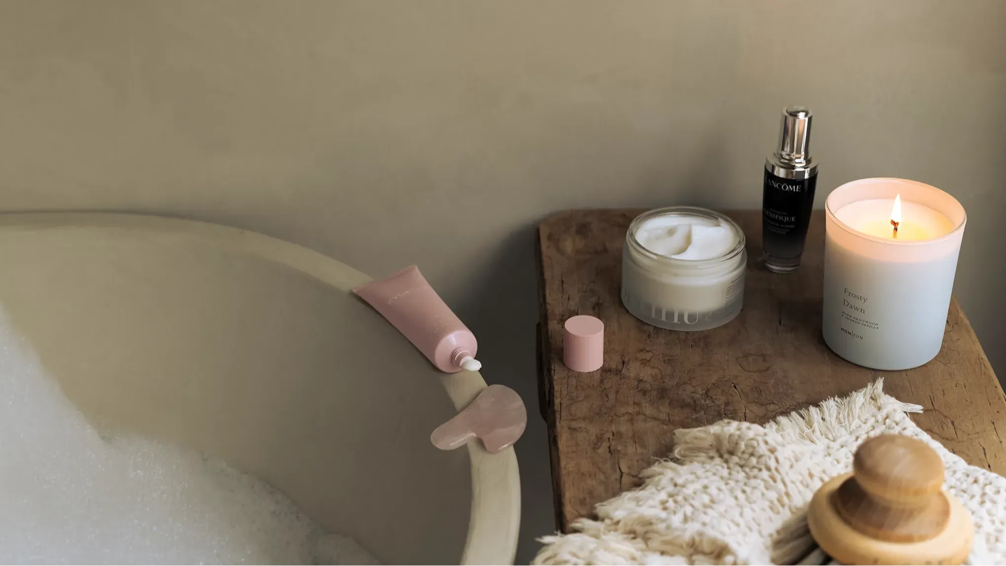Hjemmespa-stemning med badekar, hudpleieprodukter, tent duftlys og gua sha-stein på et trebord.