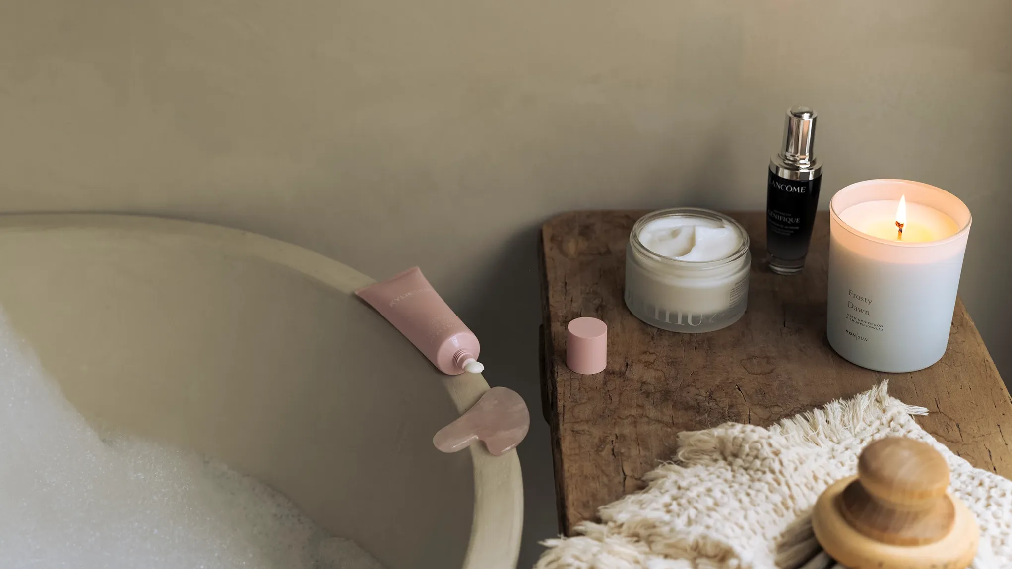 Hemmaspa-miljö med badkar, hudvårdsprodukter, tänd doftljus och en gua sha-sten på ett träbord.