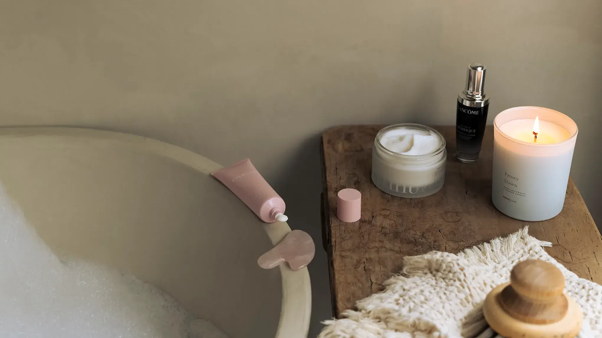 Hemmaspa-miljö med badkar, hudvårdsprodukter, tänd doftljus och en gua sha-sten på ett träbord.