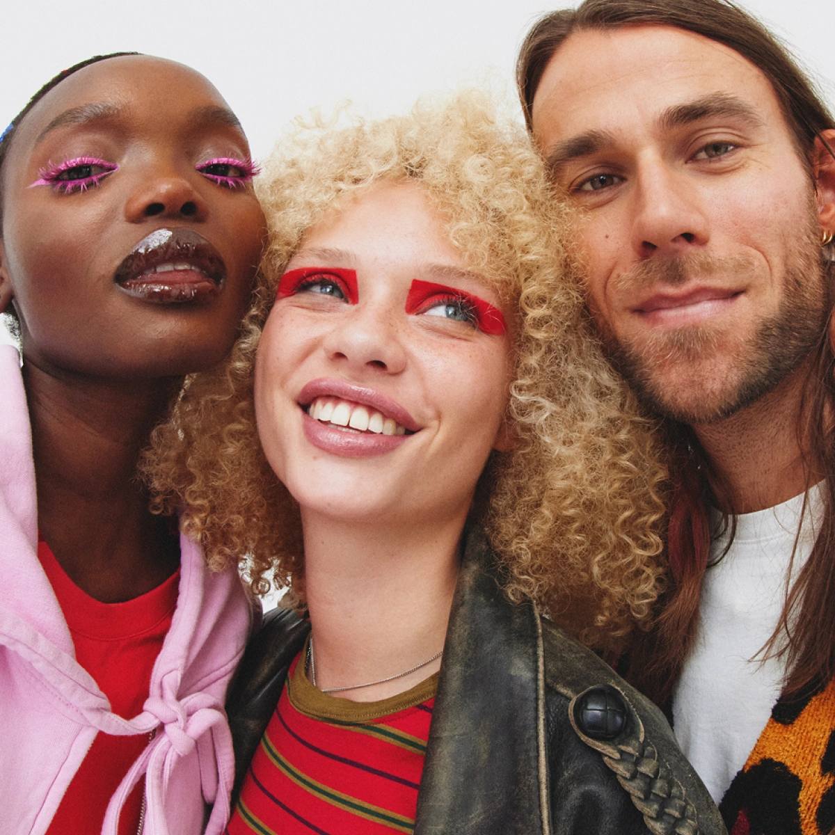 Tre personer poserar nära varandra med färgstark makeup, inklusive rosa och röd ögonskugga och glossiga läppar.