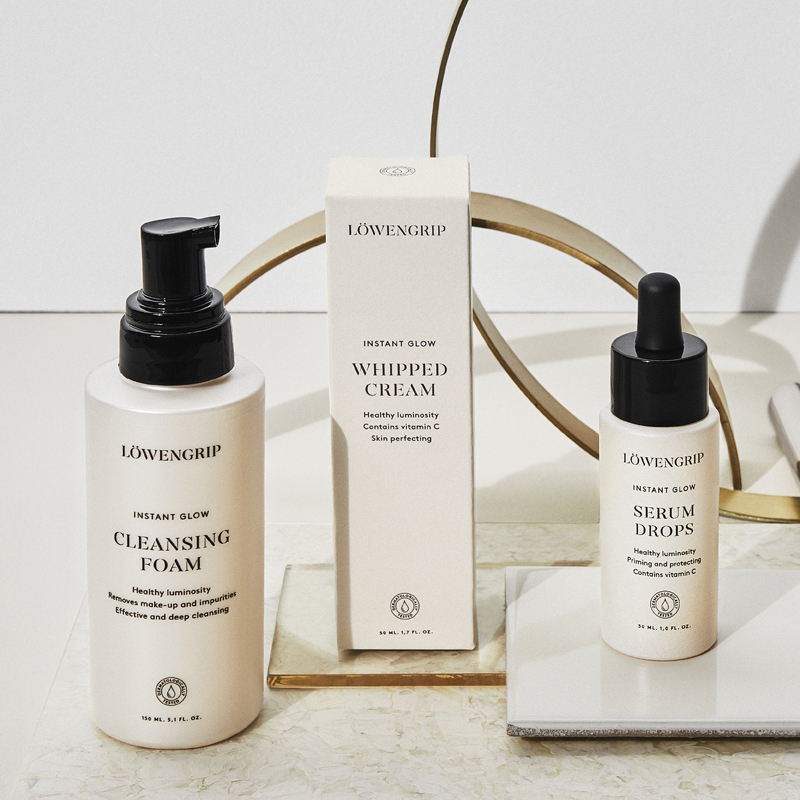 Tre hudvårdsprodukter från Löwengrips Instant Glow-serien står uppradade mot en ljus bakgrund. Alla produkter har beige förpackningar med svart text och svarta lock. Produkterna är placerade på olika plattor och en dekorativ guldring syns i bakgrunden.