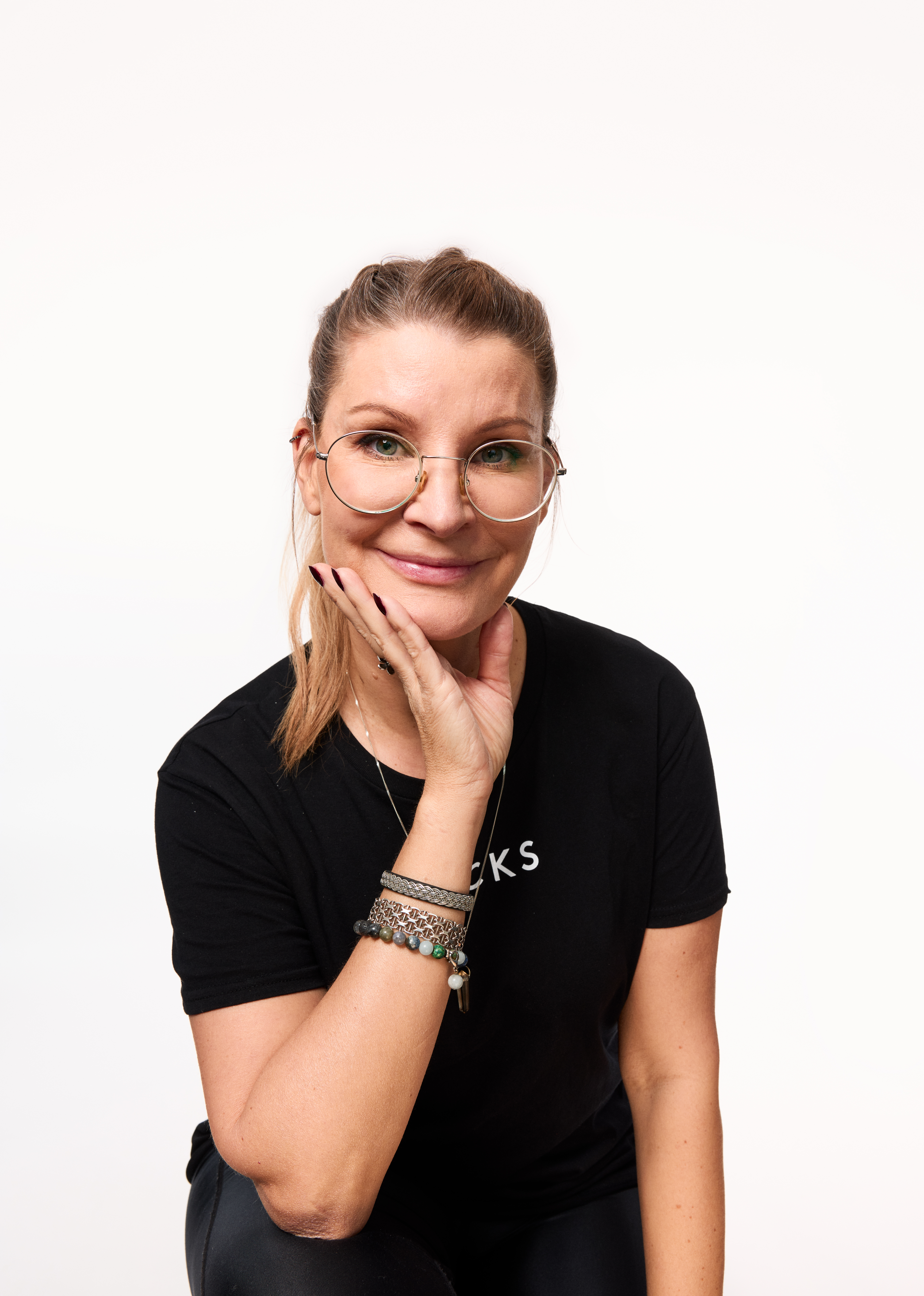 Person sitter mot vit bakgrund och tittar mot kameran. Personen har uppsatt hår, runda glasögon och ett svart t-shirt med texten ”KICKS”. Ena handen vilar mot kinden och flera armband syns på handleden.