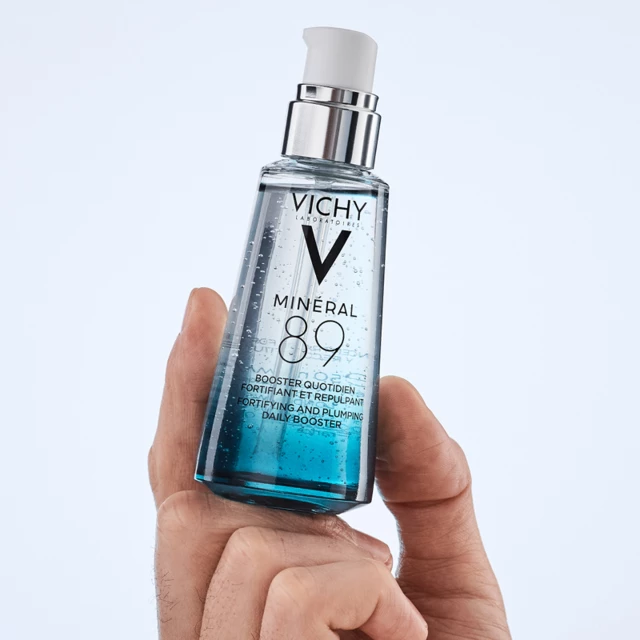 En hand håller en Vichy-produkt framför en blå vägg.