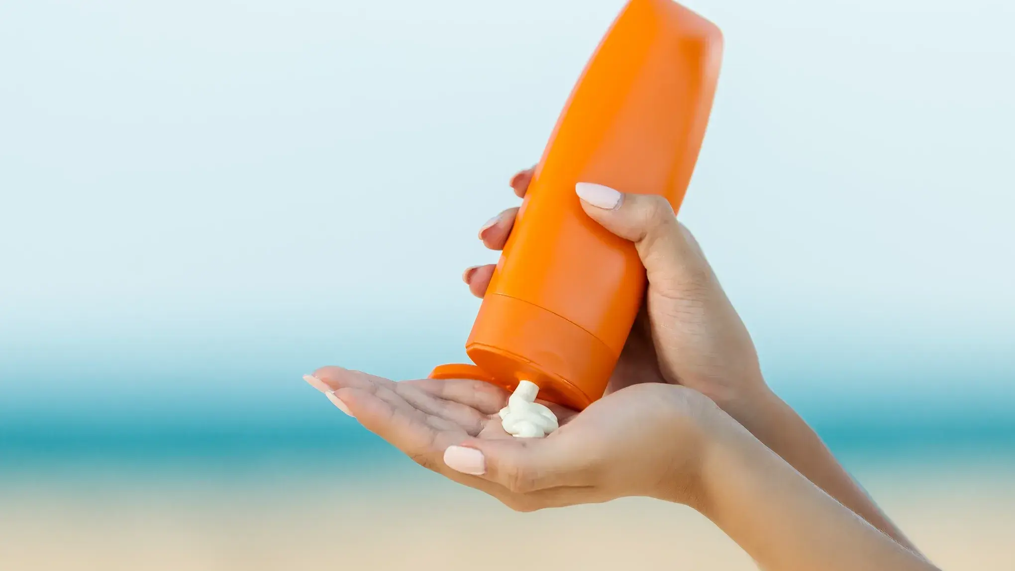 En person håller en orange flaska och applicerar solkräm på sin hand vid stranden.