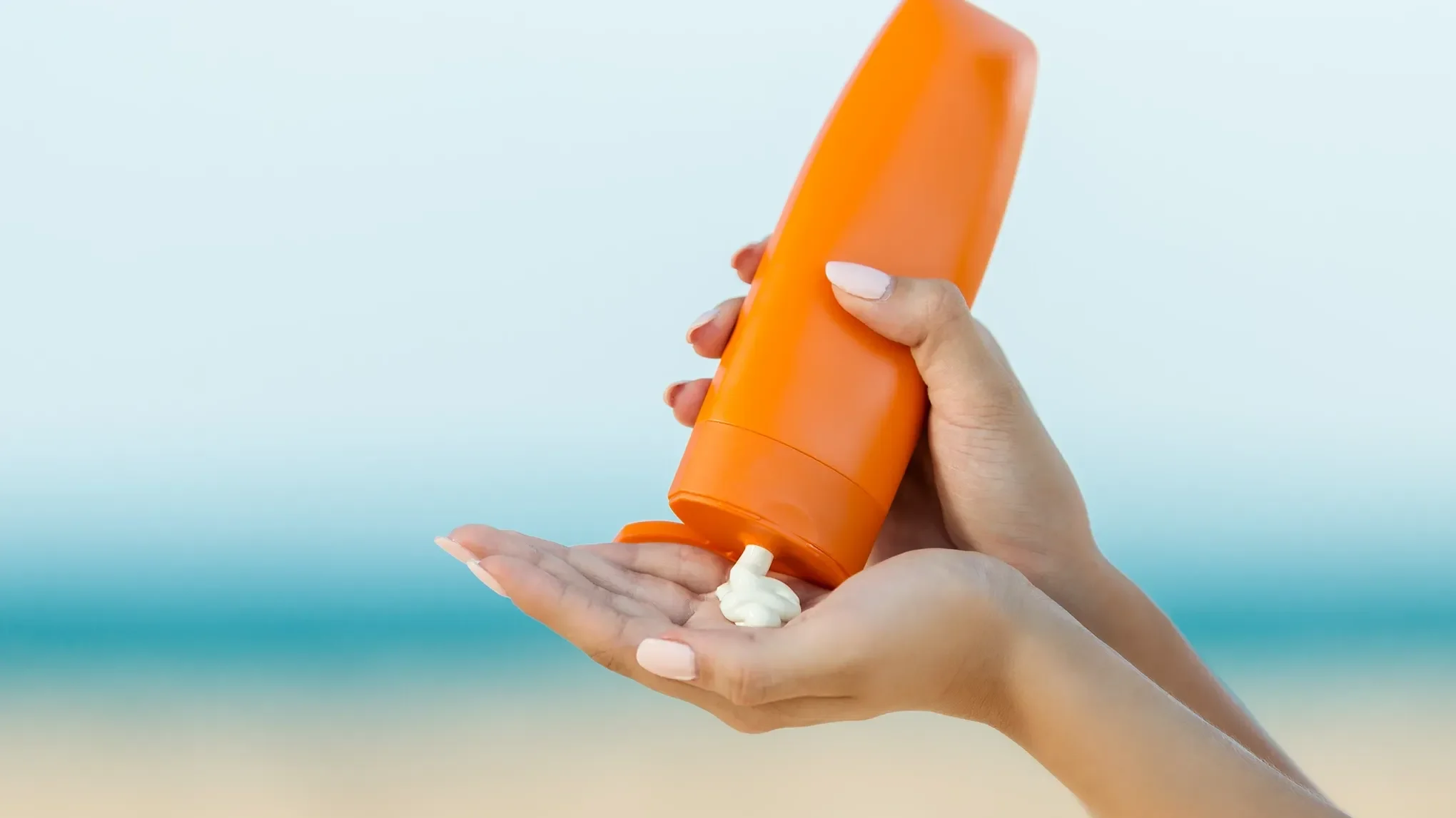 En person håller en orange flaska och applicerar solkräm på sin hand vid stranden.