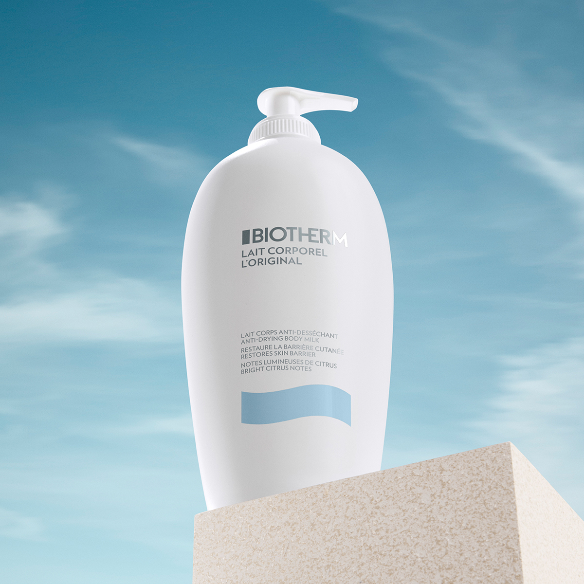 Biotherm Lait Corporel L'Original. Vartalomaito on valkoisessa pumppupullossa, jonka etupuolella on teksti ”Biotherm Lait Corporel L'original”. Pullo seisoo beigen värisellä alustalla sinistä taivasta vasten, jossa on joitakin valkoisia pilviä.