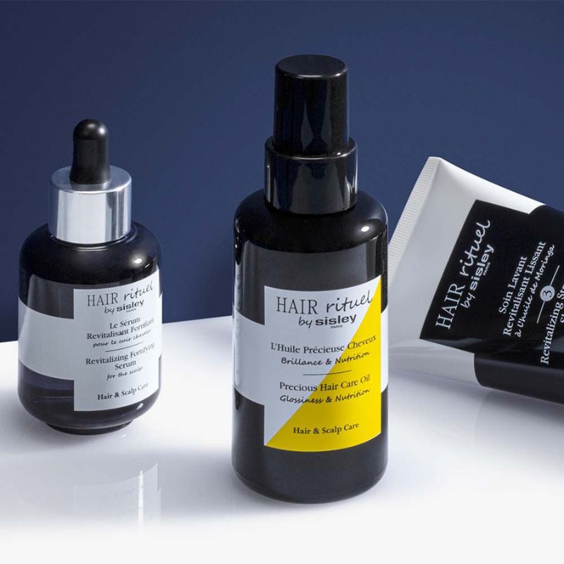 Tre hårpleieprodukter fra Sisley plassert på en hvit flate med mørkeblå bakgrunn: en flaske med pipette, en flaske med pumpe og en tube. Produktene har emballasje i svart, hvitt og gult.