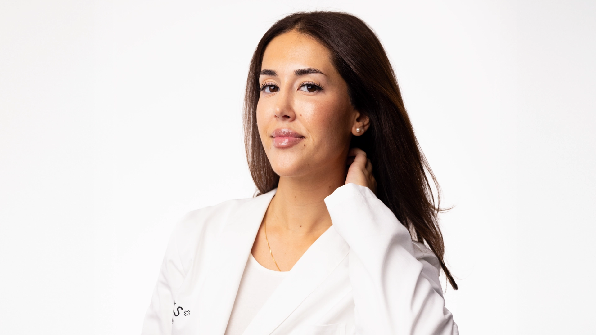 Porträtt av en person med långt, mörkt hår som bär en vit kavaj och en ljus topp. Personen står mot en vit bakgrund och håller ena handen bakom nacken.