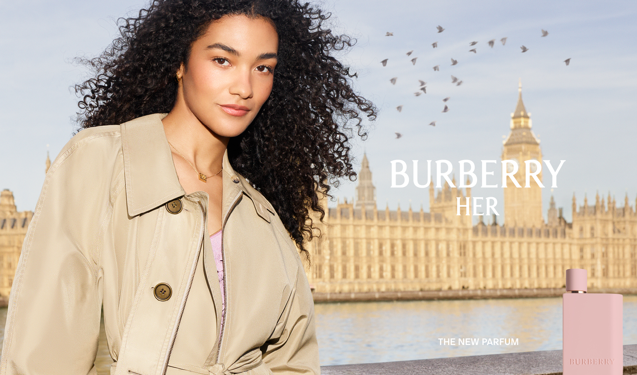 En person i trenchcoat framför en stadsmiljö med flod och byggnader, tillsammans med Burberry Her-parfym och texten “The New Parfum”.