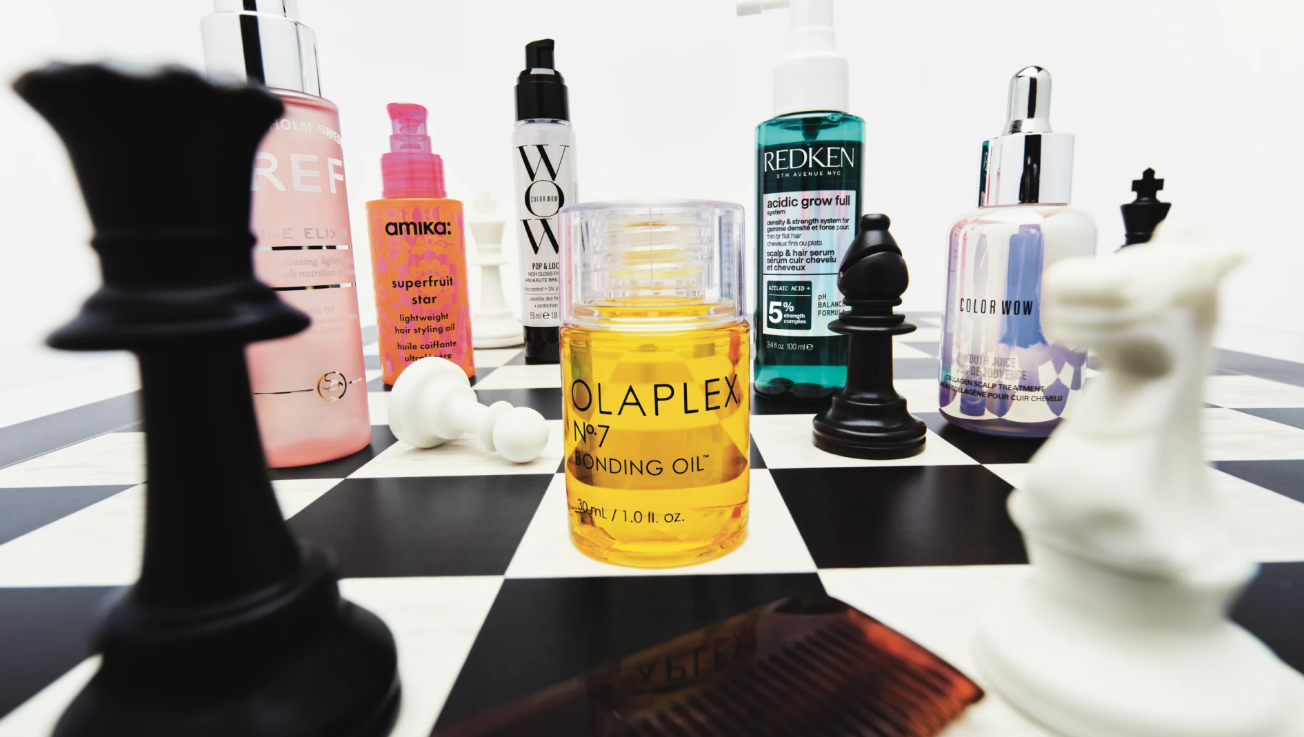 Hårpleieprodukter fra blant annet Olaplex, Redken, amika og Color Wow står blant svarte og hvite sjakkbrikker på et sjakkbrett. I midten står Olaplex No.7 Bonding Oil, 30 ml / 1.0 fl. oz.