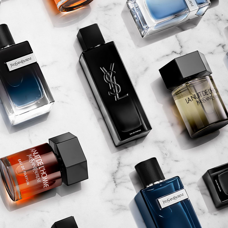 Flere parfymeflasker fra Yves Saint Laurent. Flaskene er elegant arrangert på en hvit marmorbakgrunn. Den sentrale flasken er svart med YSL-logoen i relieff. Rundt den står andre flasker i ulike farger og former, blant annet blå, brun og gjennomskinnelig.