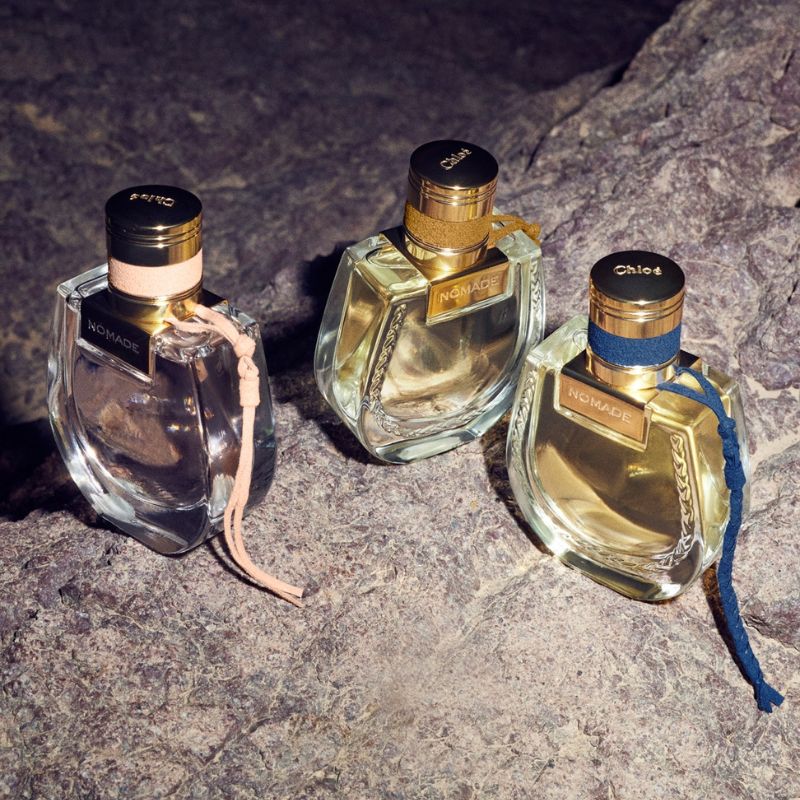 Tre parfymflaskor från Chloé med namnet "Nomade", placerade på en stenig yta. Flaskorna har guldfärgade korkar och är dekorerade med band i olika färger: rosa, beige och blått. Bakgrunden är mörk och stenig.