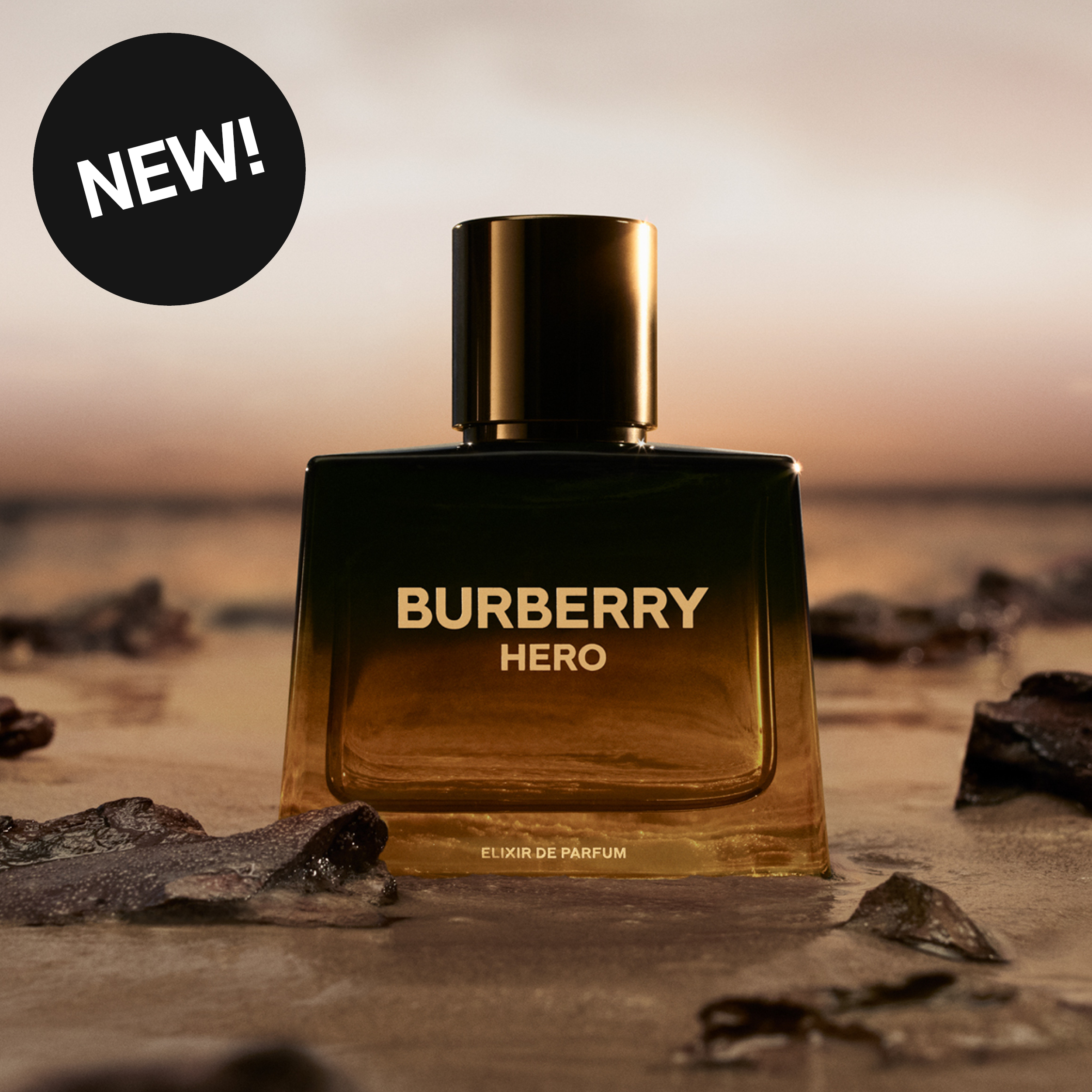 Parfyymipullo tekstillä “BURBERRY HERO ELIXIR DE PARFUM” tummassa lasipullossa kultaisella korkilla. Parfyymi on asetettu hiekalle ja kivien viereen veden äärelle auringonlaskun valossa sekä pyöreä merkki tekstillä “NEW!”.