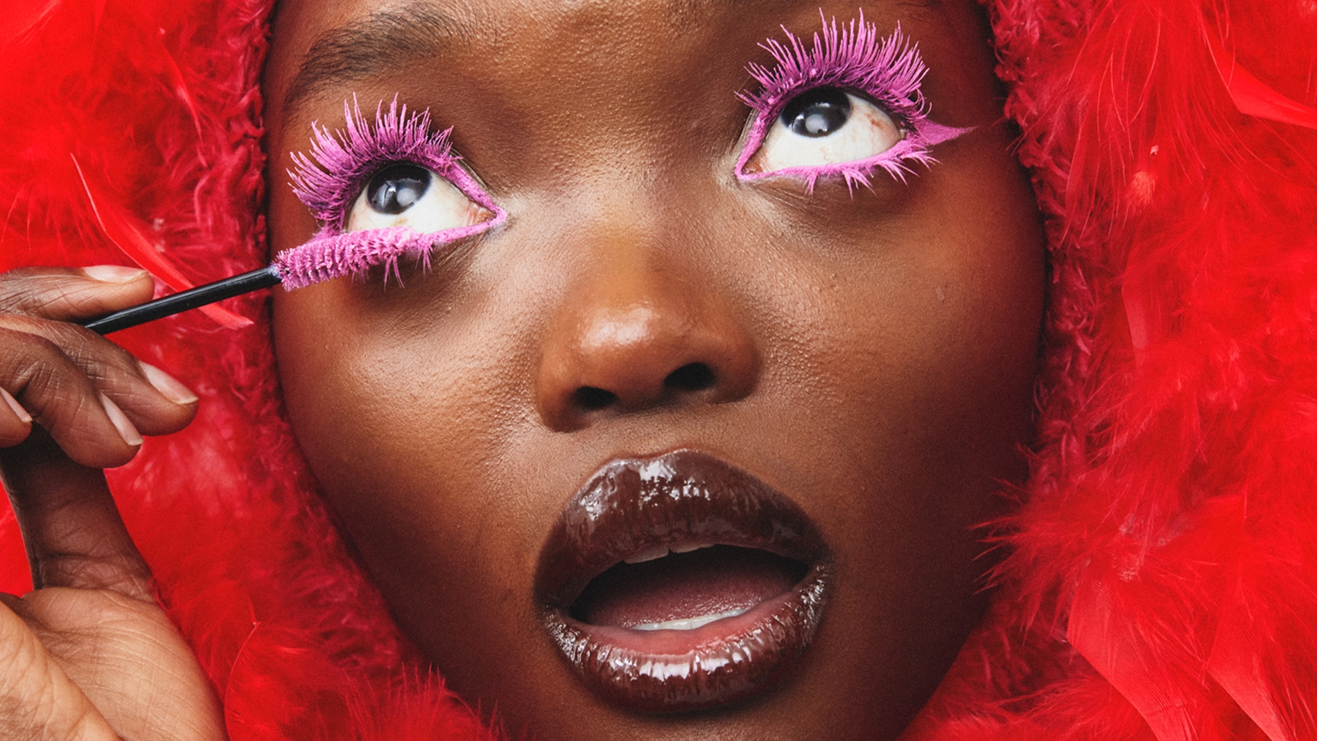 Närbild av en person som applicerar rosa mascara på sina övre fransar med en mascaraborste. Dramatiskt ögonmakeup-look med rosa fransar, glansigt läppglans och en röd fjäderhuva runt ansiktet.