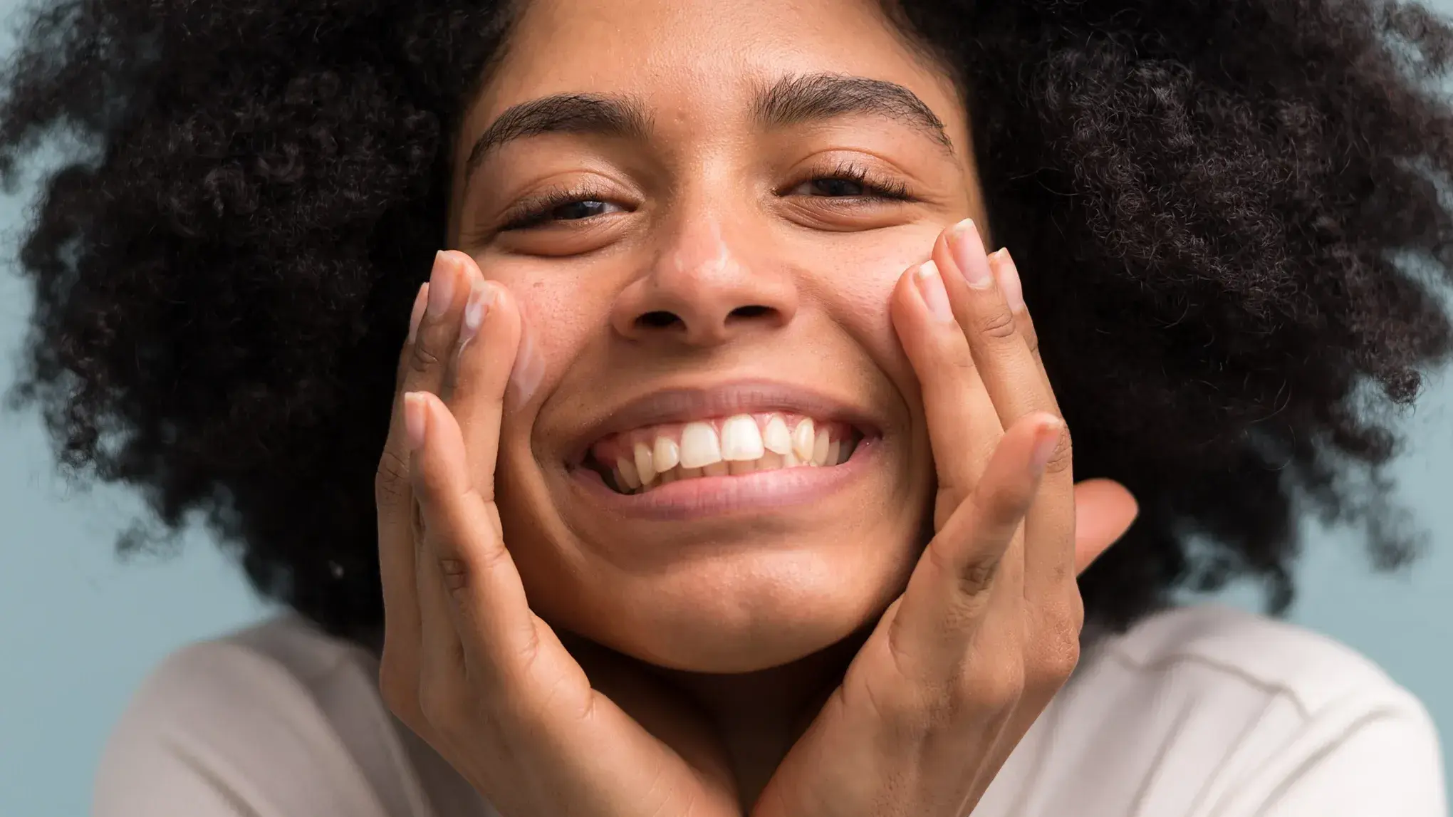 Smilende person med krøllete hår som påfører ansiktskrem.