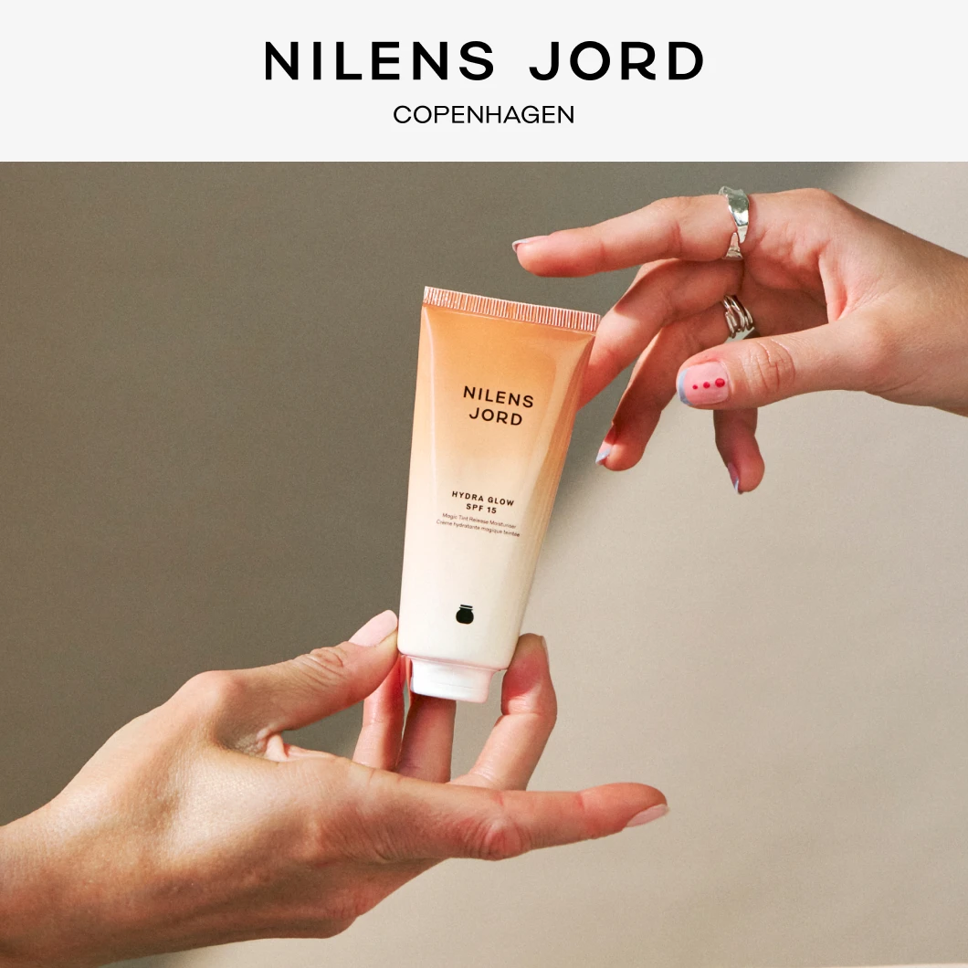 Kaksi kättä pitelee Nilens Jord -tuotetta, jossa lukee "Hydra Glow SPF 15". Yläosassa lukee "NILENS JORD COPENHAGEN".