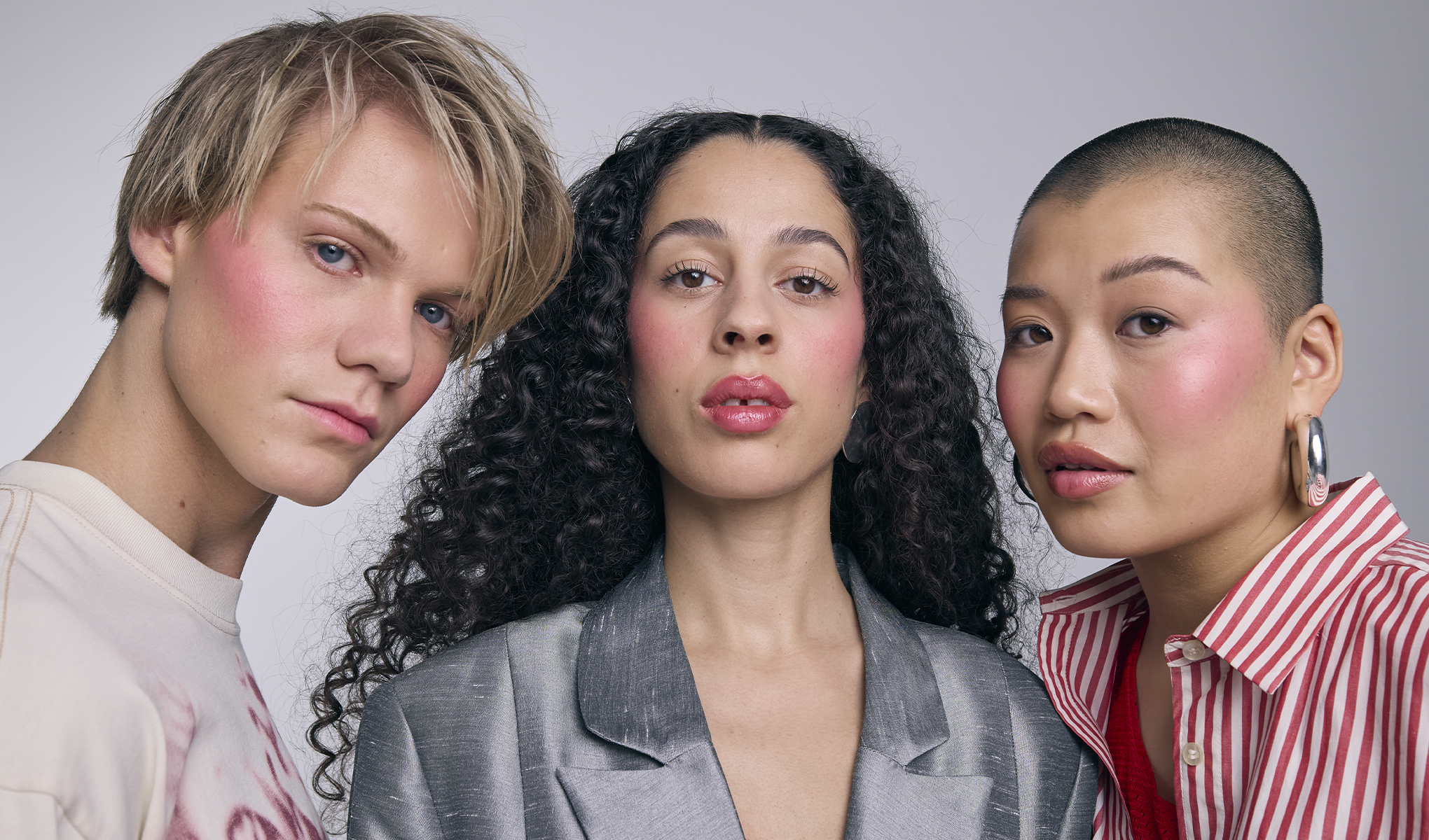 Tre personer poserar tillsammans med rosig makeup på kinder och läppar mot ljus bakgrund. Fokus på naturlig bas, blush och glansiga läppar.