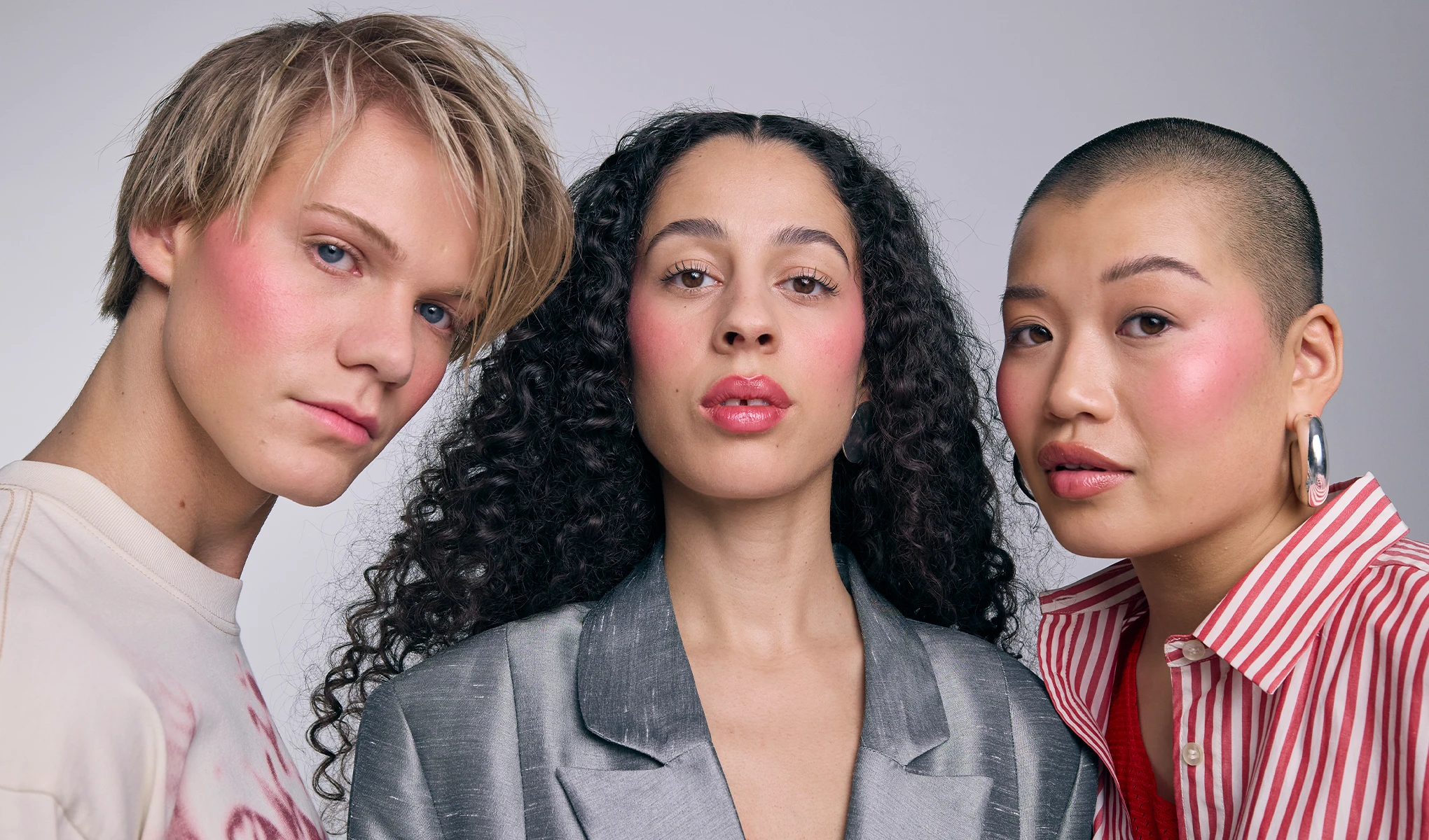 Tre personer poserar tillsammans med rosig makeup på kinder och läppar mot ljus bakgrund. Fokus på naturlig bas, blush och glansiga läppar.