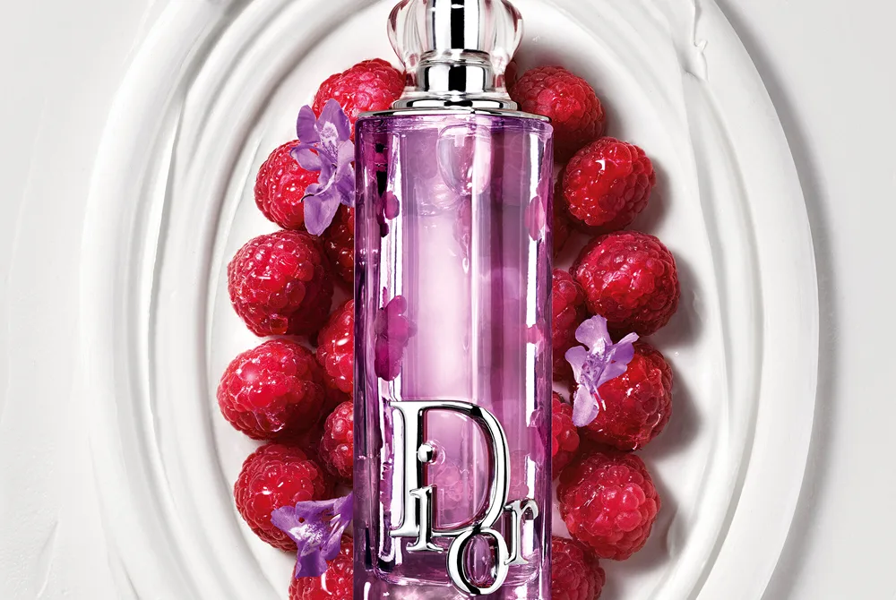 Dior Addict Lip Glow Oil i lila flaska placerad på ett dekorativt vitt fat omgivet av hallon och små lila blommor.