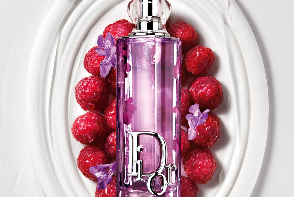 Dior Addict Lip Glow Oil i lilla flaske plassert i et dekorativt hvitt fat omgitt av bringebær og små lilla blomster.