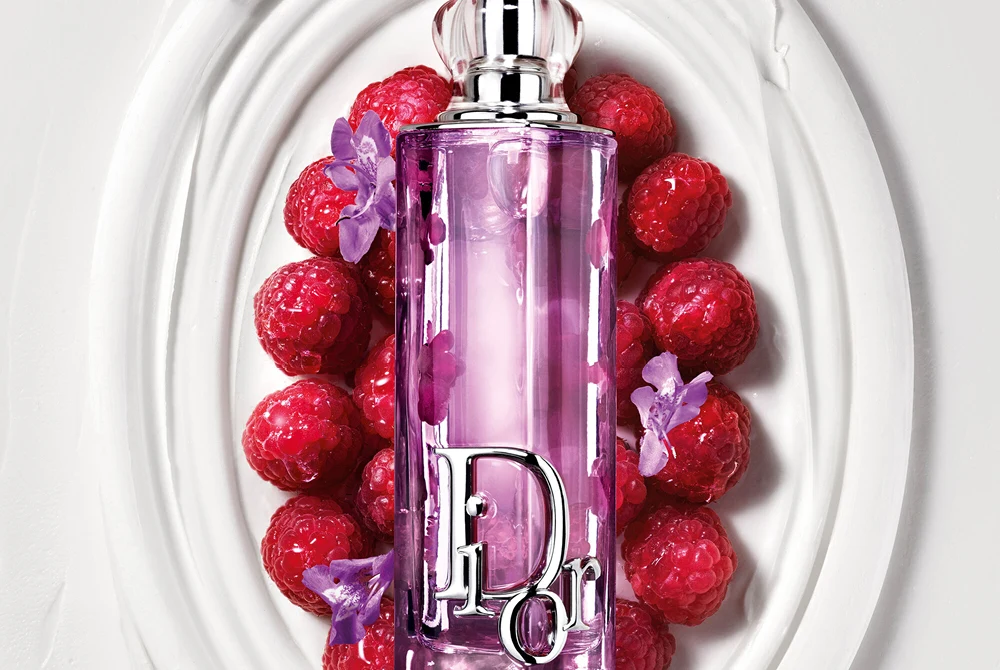 Violetti Dior Addict Lip Glow Oil -huuliöljypullo valkoisella koristeellisella vadilla vadelmien ja pienten violettien kukkien ympäröimänä.