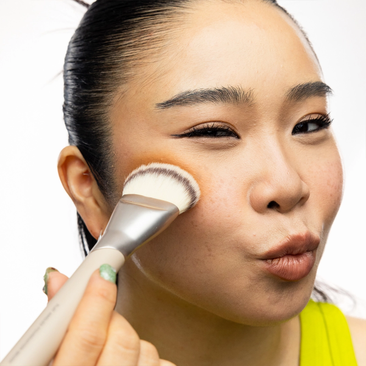 Närbild på person som applicerar contour med en tät sminkborste längs kinden. Personen blinkar med ena ögat och har ljusgrönt nagellack.