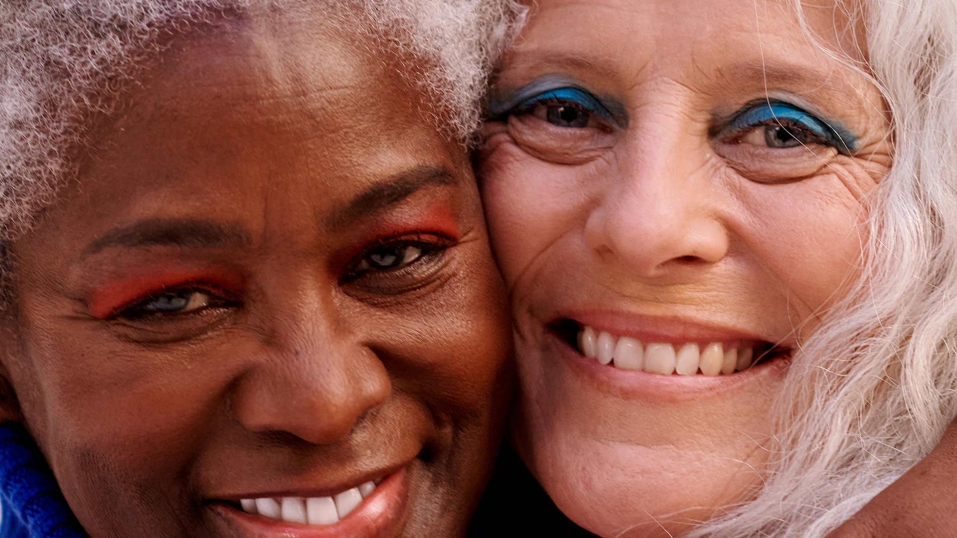 Närbild av två äldre personer som ler mot kameran med kinderna tätt intill varandra. Båda har ljus ögonmakeup i blå respektive orange nyans och grått hår. Bilden är beskuren så att endast ansikten och delar av håret syns.