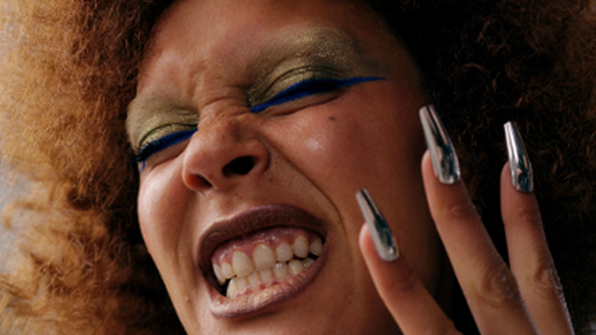 Närbild av en person som ler brett mot kameran. Personen har lockigt hår, grafisk ögonmakeup med färgade linjer, markerade ögonfransar och långa, blanka naglar som hålls upp nära ansiktet.