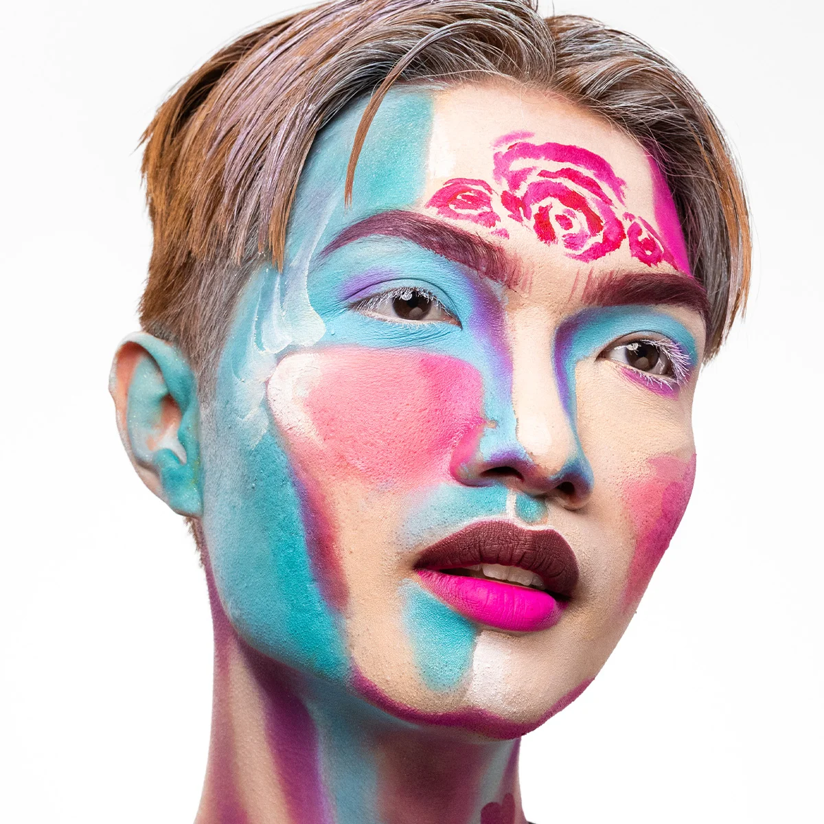 Närbild av kreativ ansiktsmålning i blå, rosa, lila och beige toner, med målade rosor i pannan, markerade bryn och tvåfärgade läppar som skapar ett konstnärligt helhetsintryck.