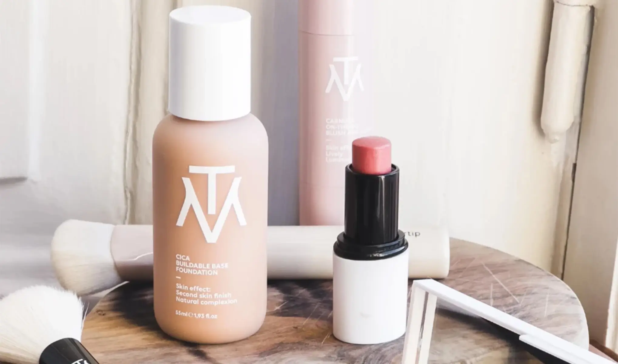 En närbild av olika kosmetikprodukter på ett bord, inklusive en foundationflaska, en läppstiftsapplikator, och en borste. Produkterna har en stilren design med vita och rosa färger och är märkta med logotypen "MTM".