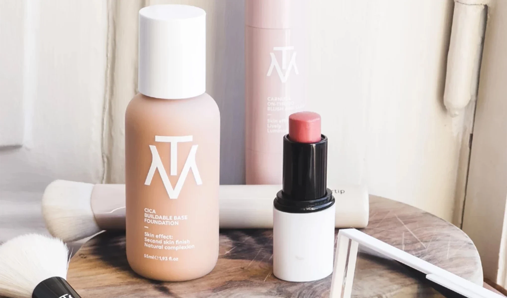 En närbild av olika kosmetikprodukter på ett bord, inklusive en foundationflaska, en läppstiftsapplikator, och en borste. Produkterna har en stilren design med vita och rosa färger och är märkta med logotypen "MTM".