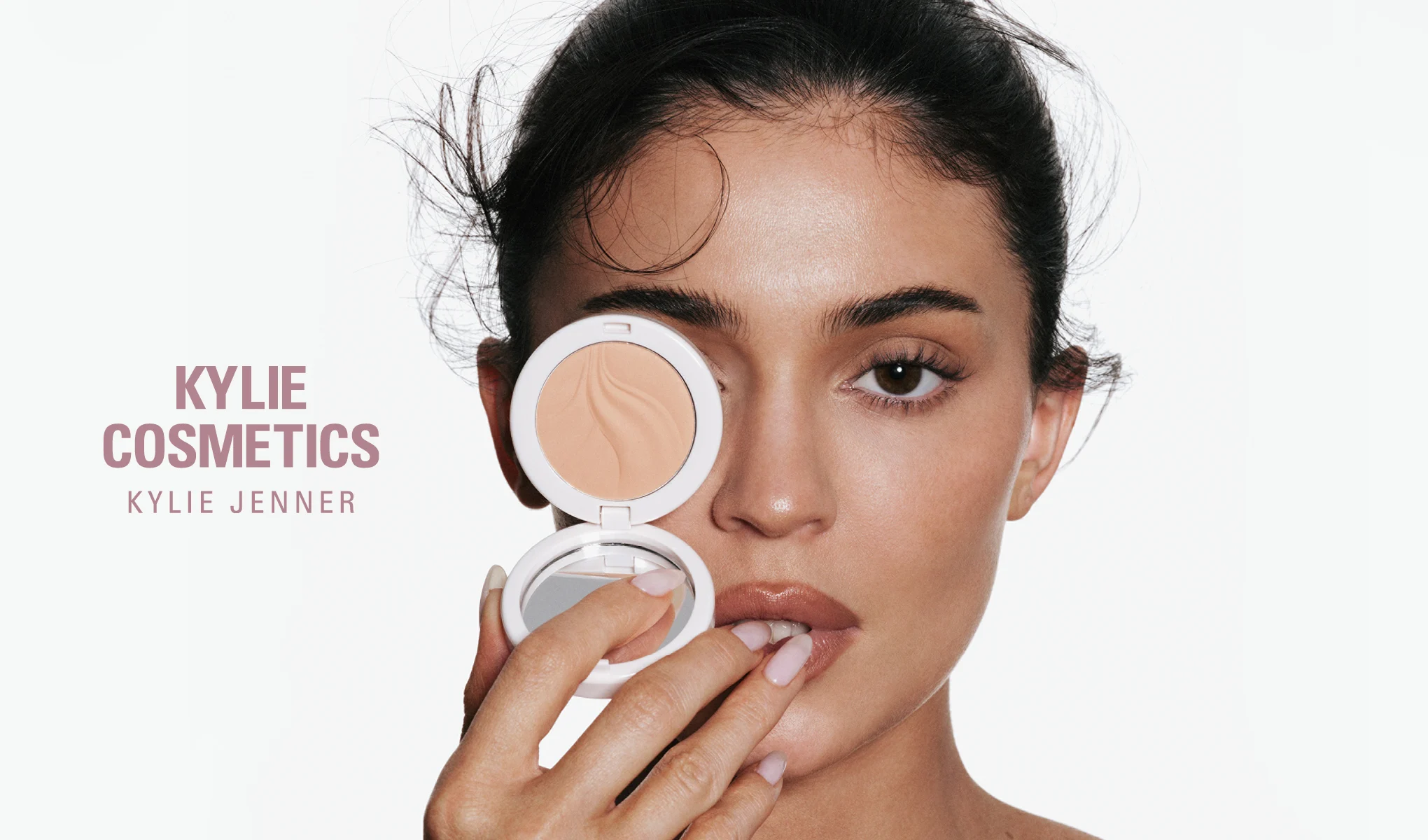 En person håller upp en öppen Kylie Cosmetics-kompakt med puder framför ena ögat. Synlig text: "KYLIE COSMETICS" och "KYLIE JENNER".
