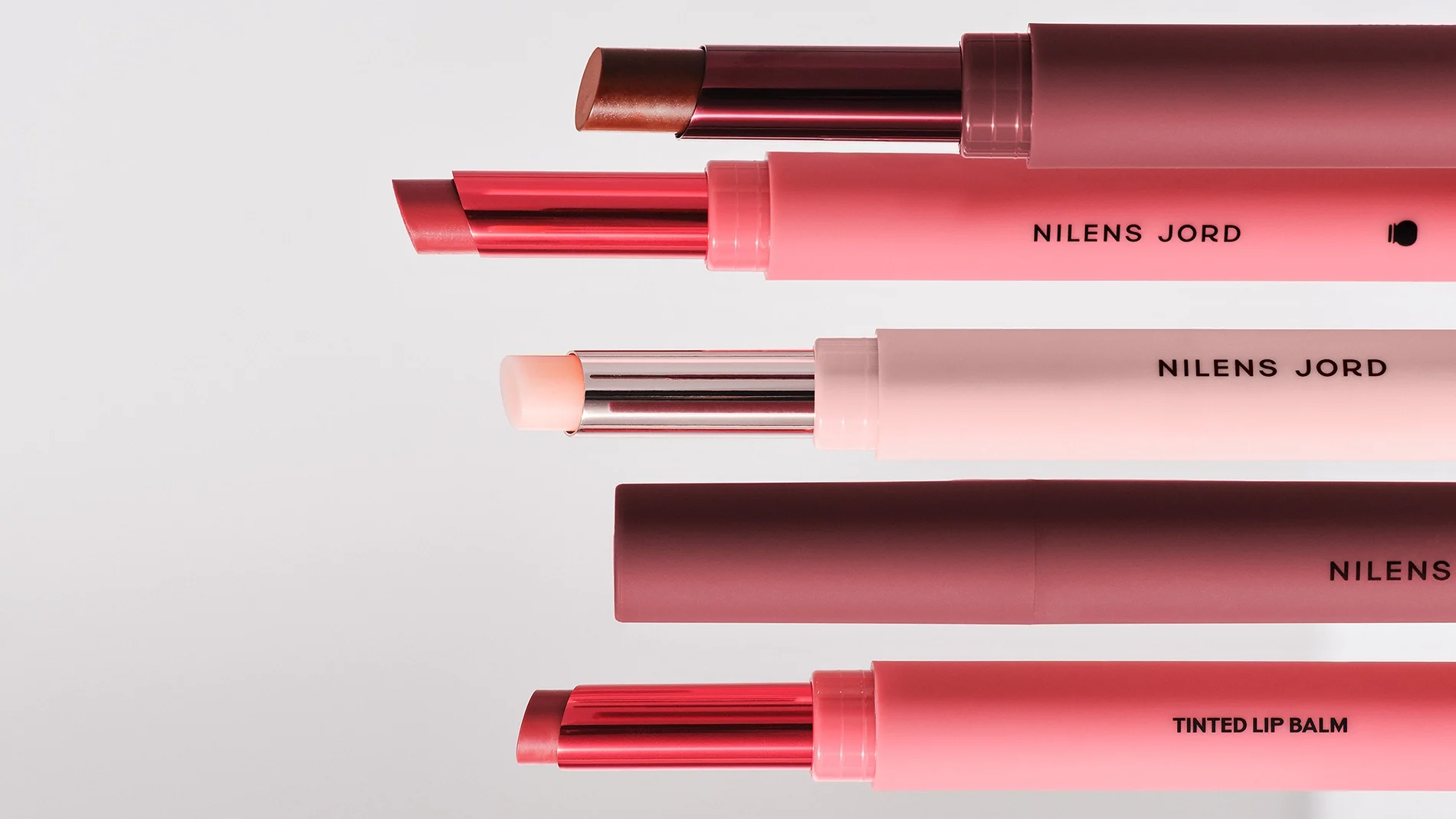 Fem leppebalmer fra Nilens Jord i rosa og vinrød emballasje mot lys bakgrunn, med synlig tekst "TINTED LIP BALM", "NILENS" og "NILENS JORD". Leppebalmene vises i rosa, transparent lys og brun nyanse.