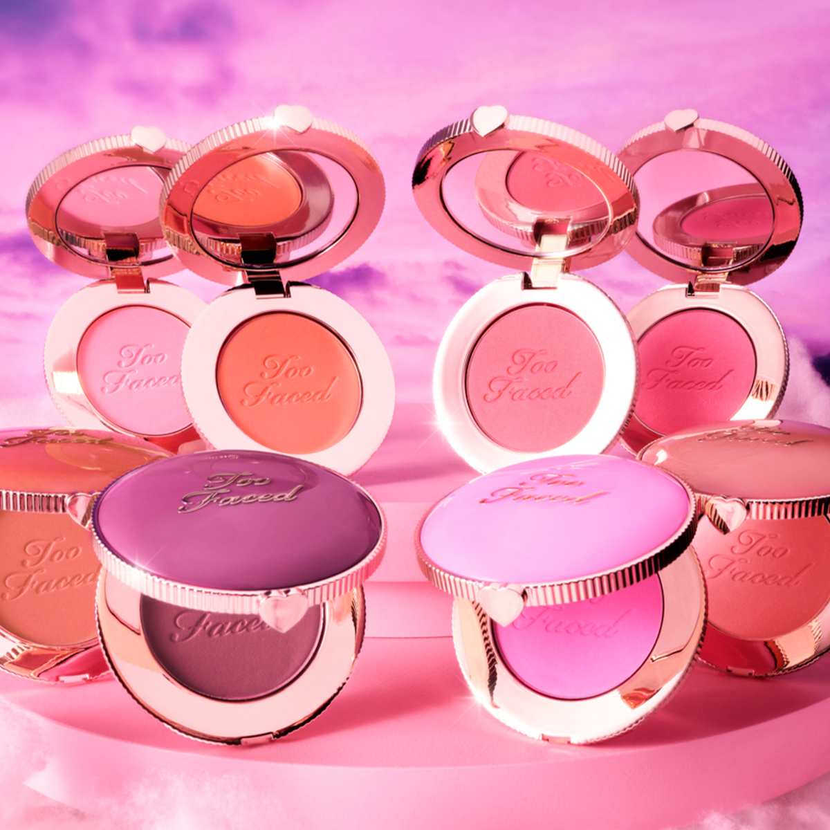 Too Faced-blushprodukter som står på en lys rosa pall med en rosa bakgrunn. Blushene har forskjellige nyanser og emballasjen har en hjertepynt.