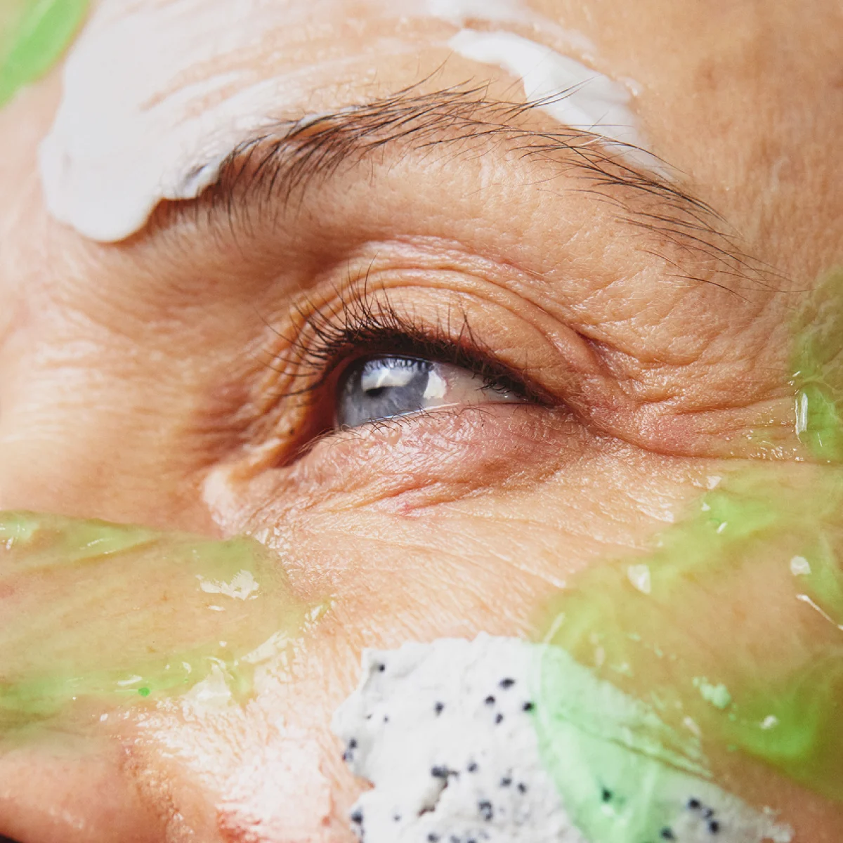 Närbild av ett öga och en del av ansiktet på en person med olika hudvårdsprodukter applicerade på huden, bland annat en grön gel och en vit exfolierande kräm.