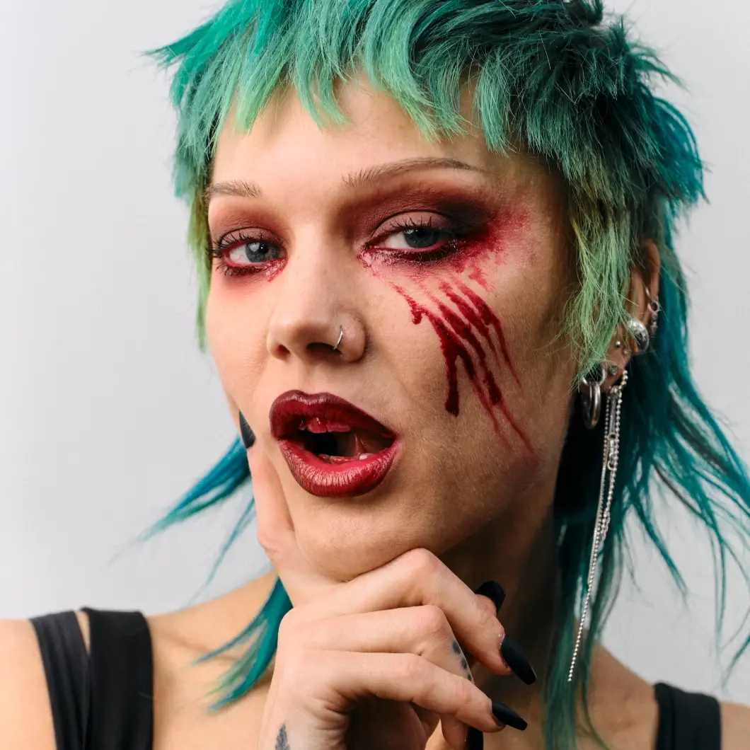 Henkilöllä on turkoosit hiukset ja näyttävä Halloween-meikki, jossa on punainen silmämeikki, punaisia raapimisjälkeä muistuttavia viivoja poskella ja tummanpunainen huulipuna.