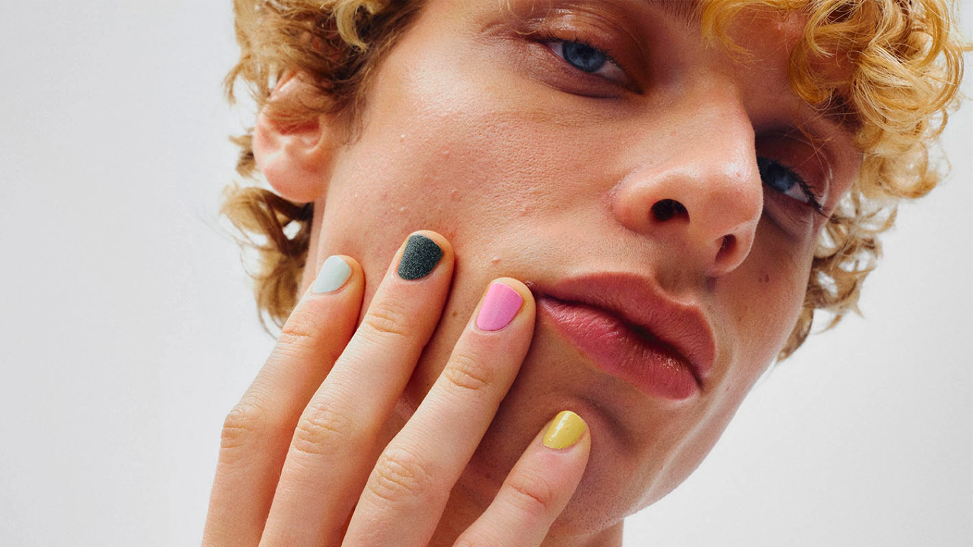Lähikuva kasvoista, käsi poskella ja kynnet lakattu eri väreillä kuten pinkki, keltainen, vihreä ja vaaleansininen. Lyhyet kiharat hiukset, luonnollinen ihon tekstuuri ja neutraali tausta.