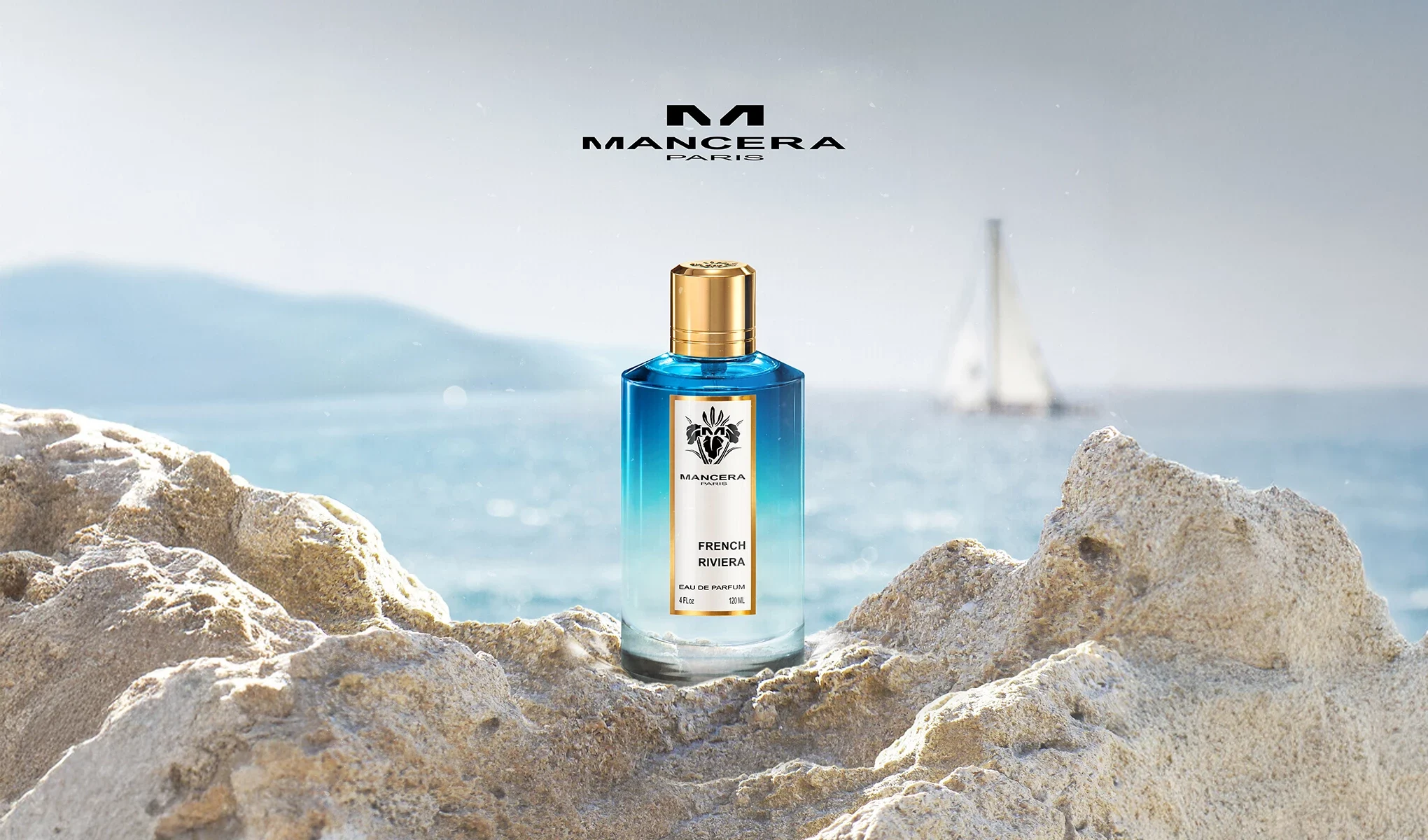 Mancera French Riviera-parfymflaska står på klippor, med havet och en segelbåt i bakgrunden.