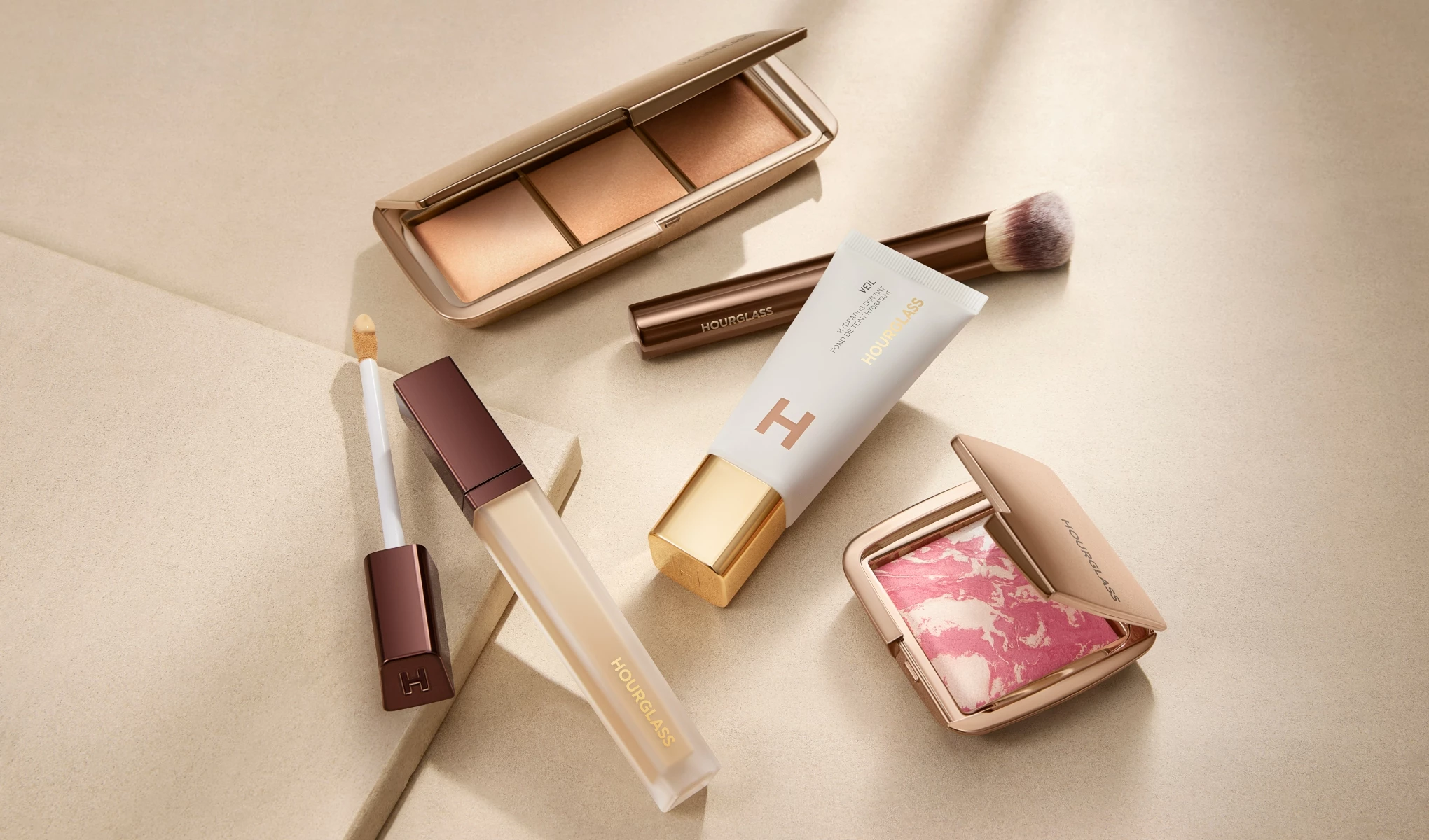 Olika kosmetikaprodukter från Hourglass, inklusive en foundation, en concealer, en puderpalett, en rouge, en sminkborste och en mascara, arrangerade på en beige yta.
