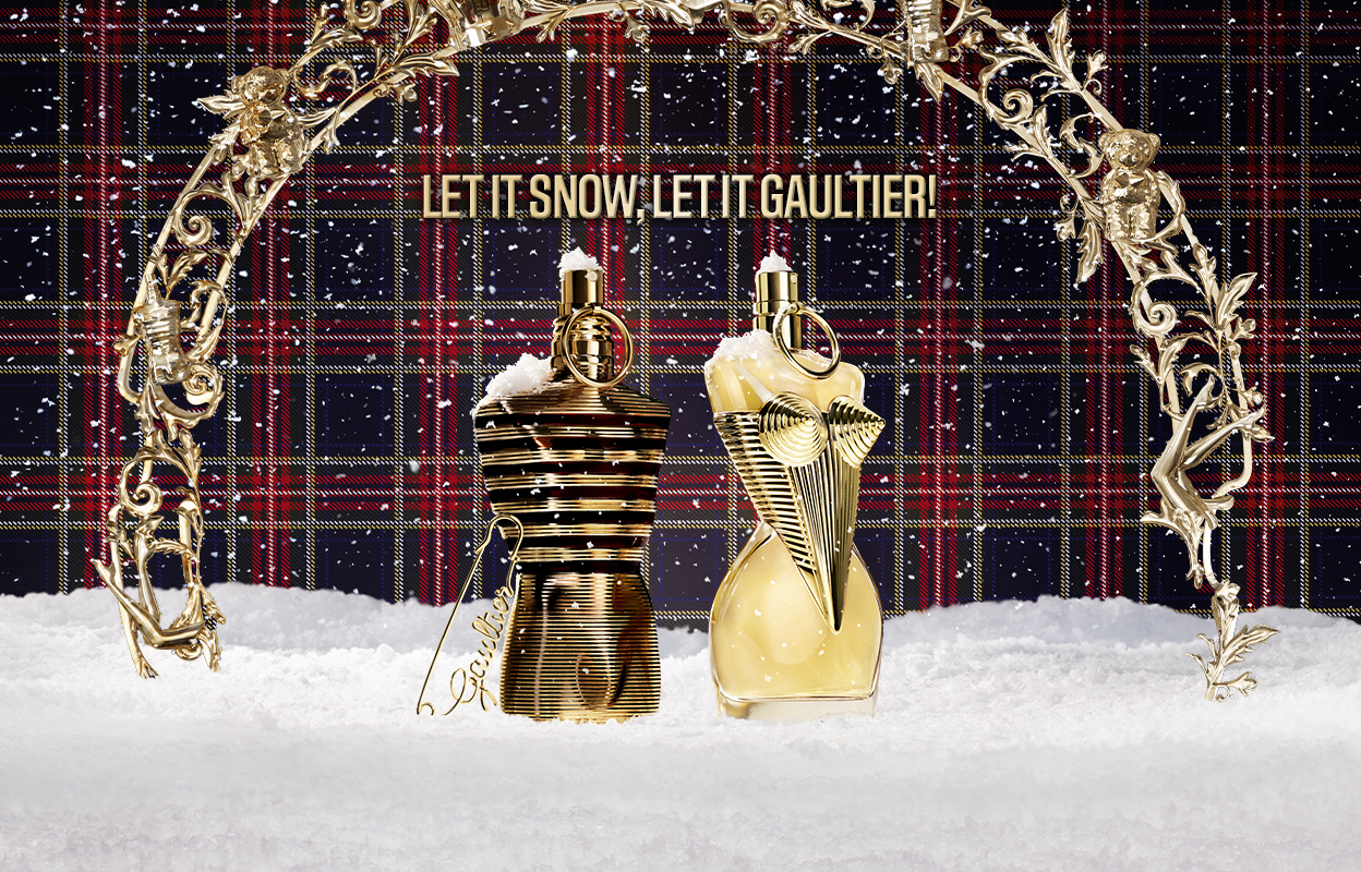 To parfymeflasker, den ene gullbelagt og den andre gyllen, står på en snødekt overflate under en dekorativ bue. Bakgrunnen har rutemønster og teksten "LET IT SNOW. LET IT GAULTIER!".