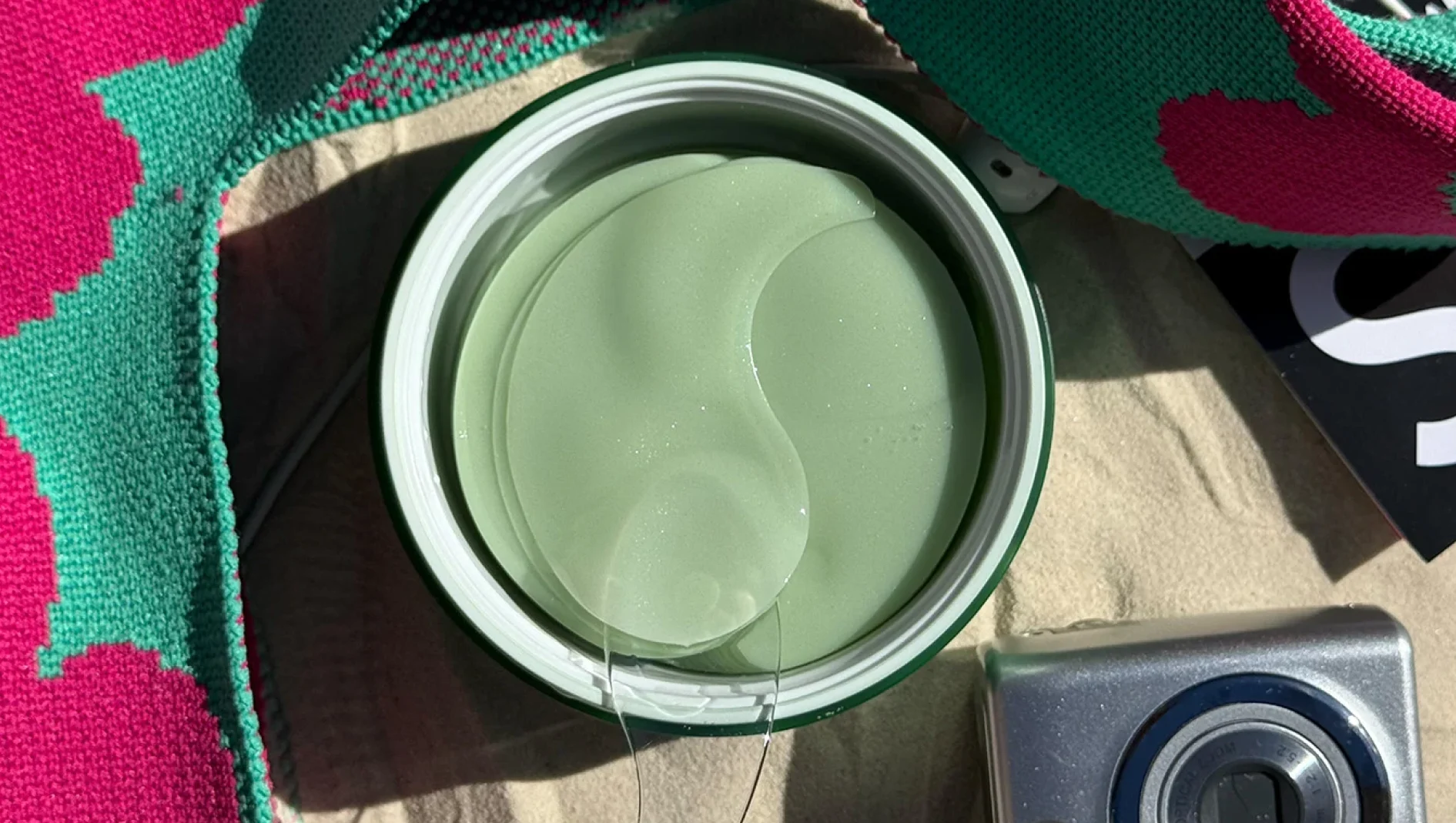 Åpen krukke med grønne øyemasker, med væske som drypper, ved siden av et rosa og turkis strikket stoff og et sølvfarget kamera.