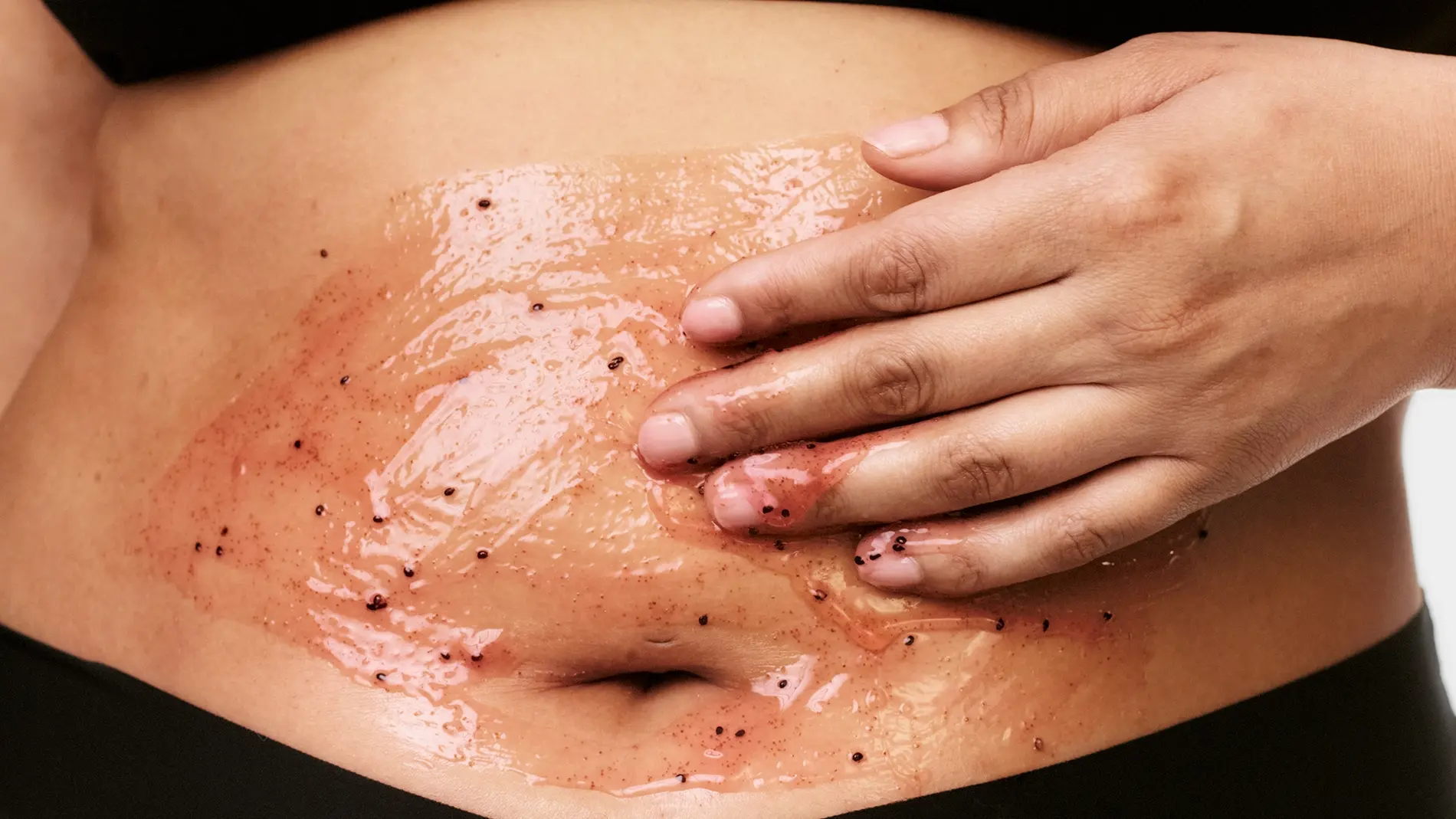 Närbild på en kroppsskrubb med geléliknande peeling och mörka exfolierande korn som appliceras på magen med handen. Hudvårdsprodukt för kroppspeeling och exfoliering med glansig finish på huden.