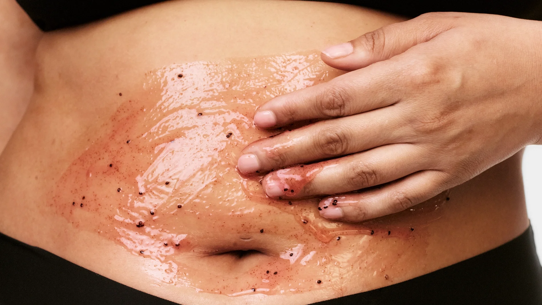 Närbild på en kroppsskrubb med geléliknande peeling och mörka exfolierande korn som appliceras på magen med handen. Hudvårdsprodukt för kroppspeeling och exfoliering med glansig finish på huden.