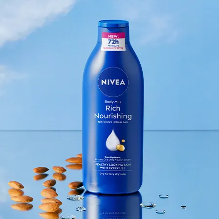 En blå flaska från Nivea placerad mot en ljusblå bakgrund. Flaskan har texten "NEW: 72h Moisture" och "Rich Nourishing" på etiketten. Runt flaskan ligger mandlar och några vattendroppar.