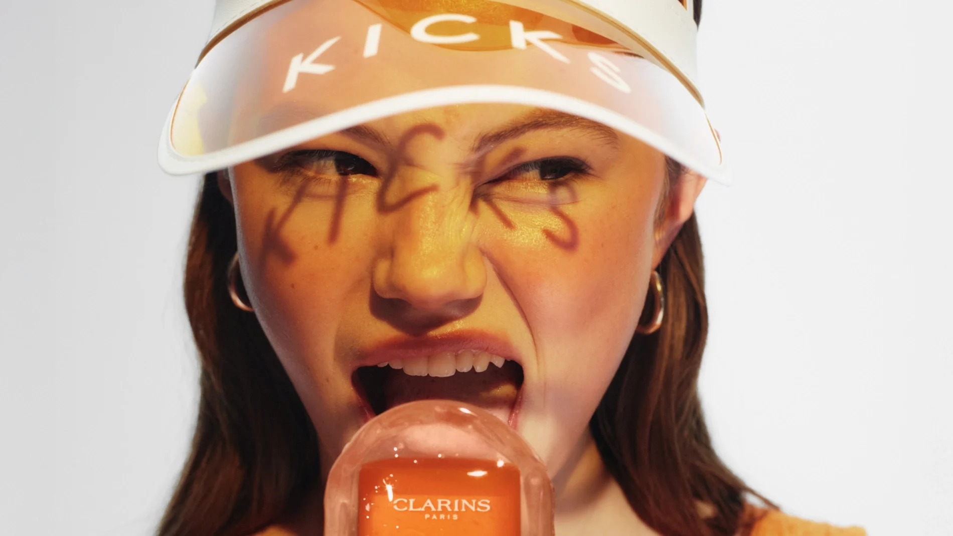 En person bär ett vitt solskärm med texten "KICKS" och håller upp en Clarins Paris Invisible Sun Care Stick High Protection SPF 50 som är utformad som en glasspinne. Ordet "KICKS" är skrivet över ansiktet, och personen poserar med öppen mun mot produkten. Orange toner och fokus på solskydd framhäver hudvård för soliga dagar.