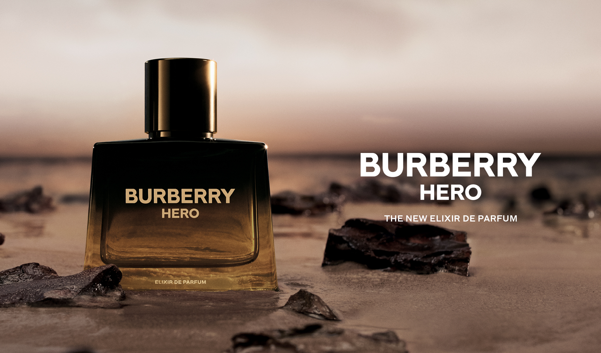 Burberry Hero Elixir de Parfum-flaske plassert i vannkanten ved solnedgang, med teksten «Burberry Hero – The New Elixir de Parfum».