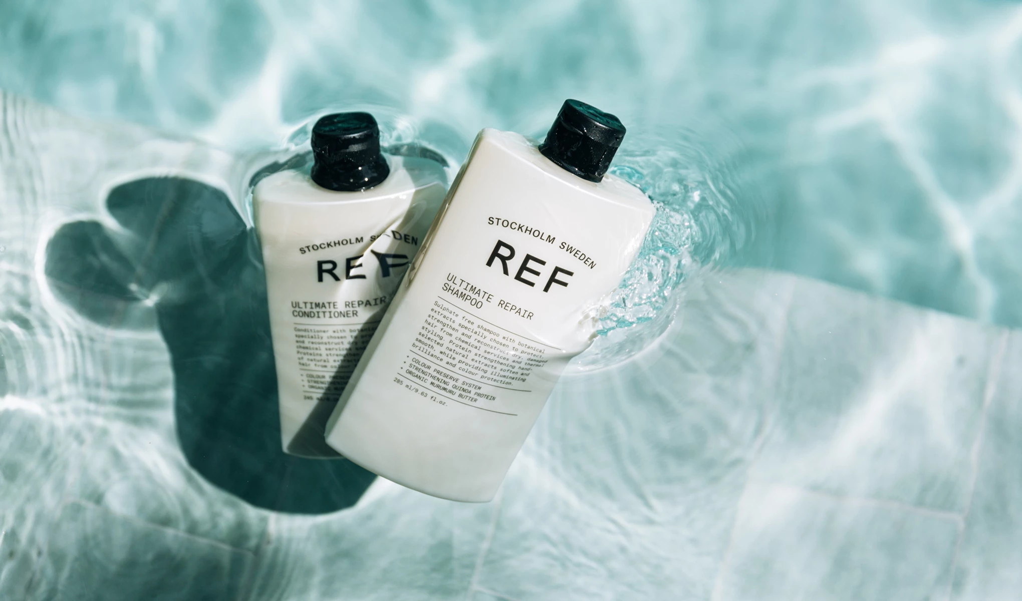 Kaksi REF Ultimate Repair -tuotetta (shampoo ja hoitoaine) kelluu kirkkaassa, sinisessä vedessä.