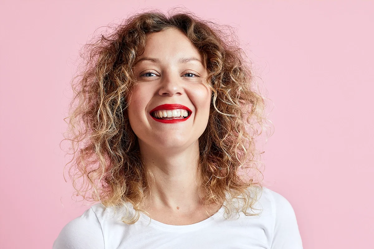 Person med lockigt hår och klarrött läppstift framför rosa bakgrund. Naturlig hudmakeup och vit topp framhäver en fräsch beautylook med fokus på läppsmink och hår.