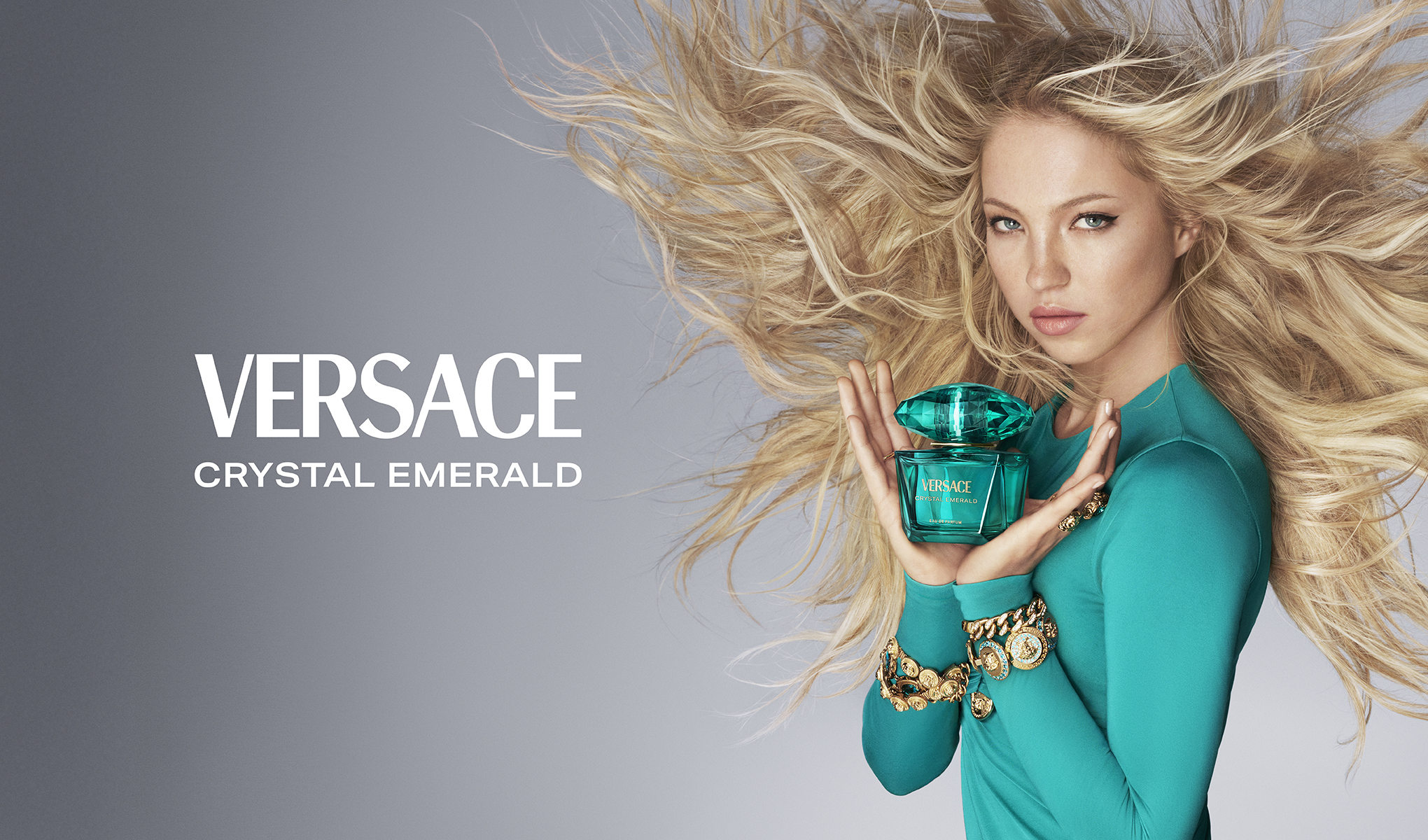 Nainen pitelee Versace Crystal Emerald -hajuvettä harmaata taustaa vasten.