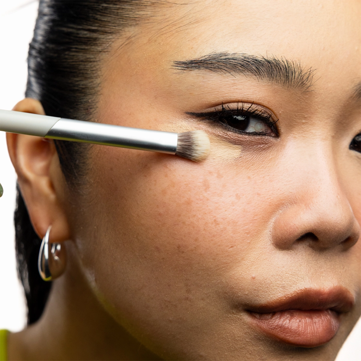Nærbilde av person som påfører concealer med en flat sminkekost under øyet. Personen bruker sølvfargede øredobber.